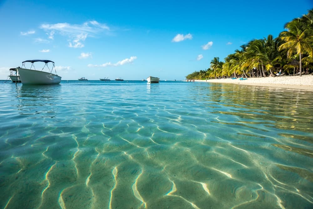 Calm, clear water reflects the blue sky and boats near a sandy beach lined with palm trees.
