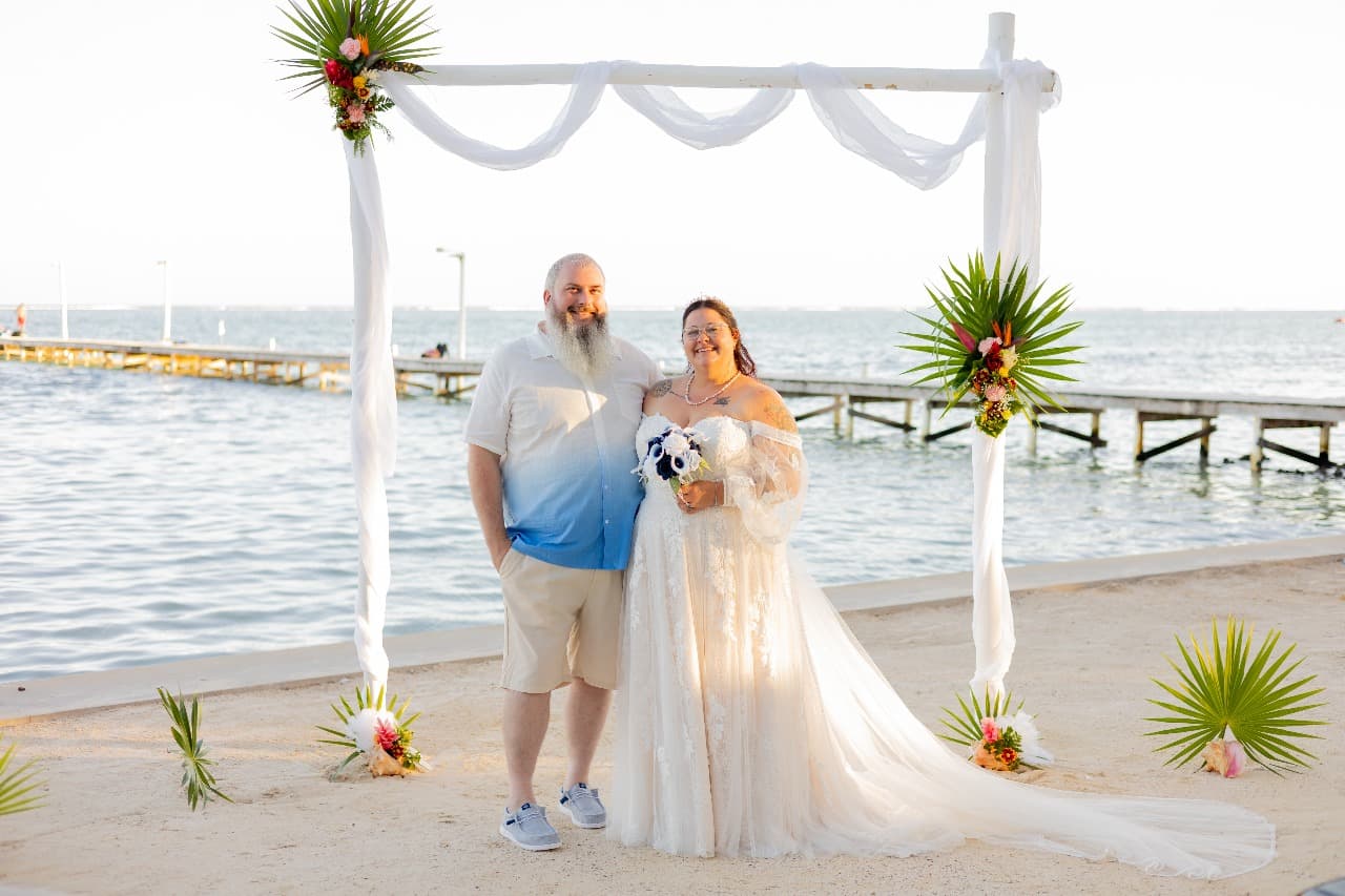 A couple stands happily together under a floral arch by the water, smiling with a beach backdrop.