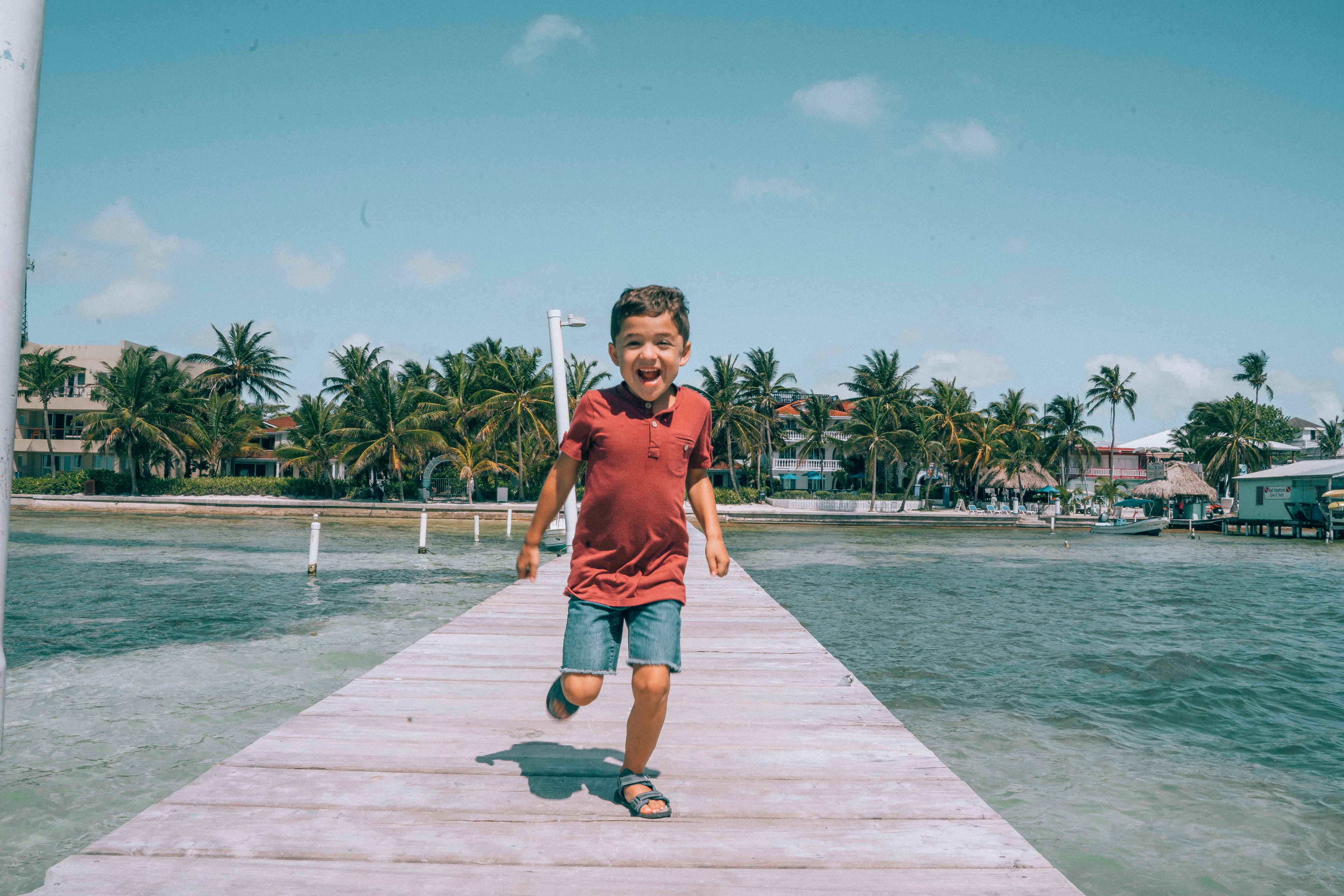 A joyful boy runs along a wooden dock surrounded by palm trees and water.