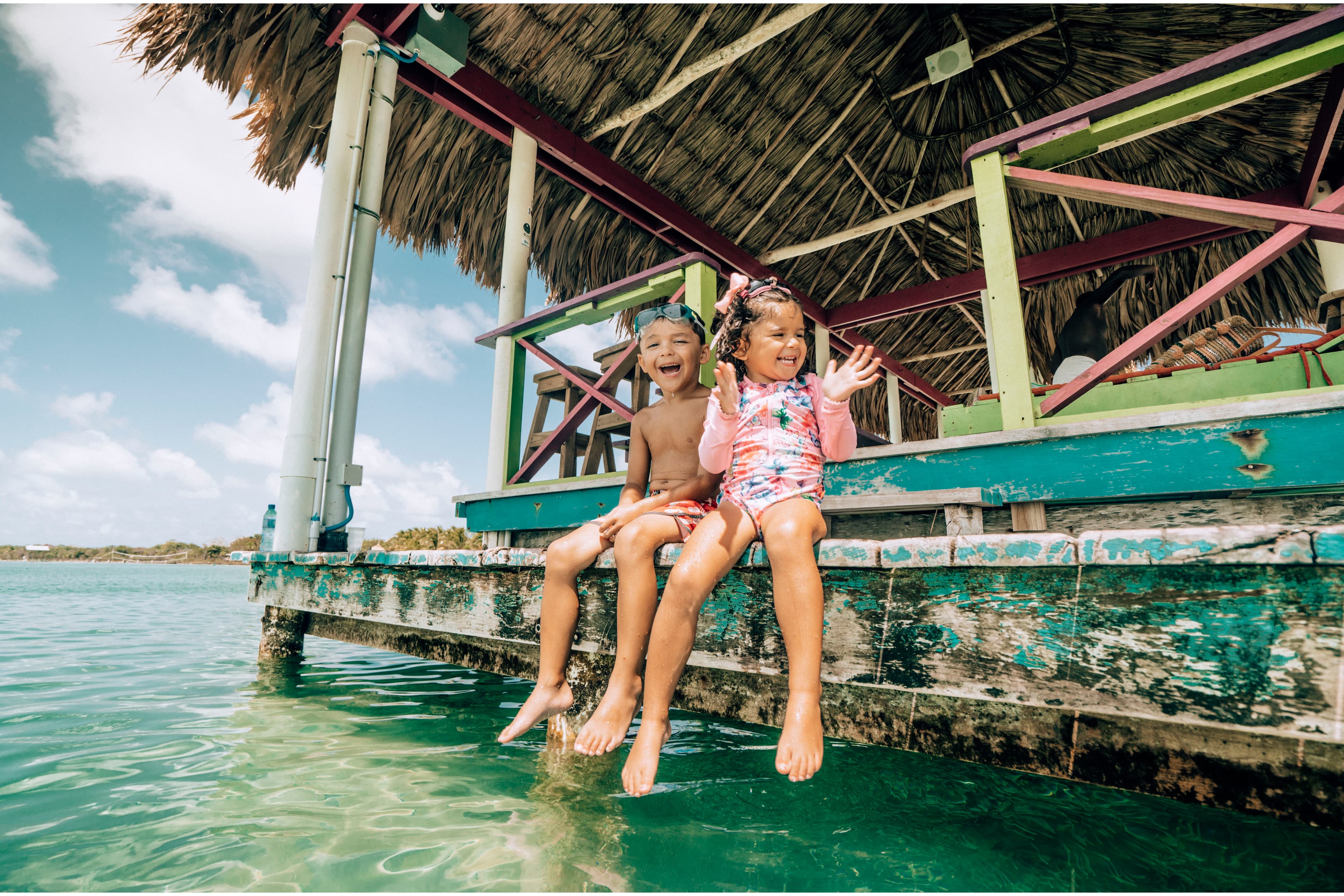 Two children sit on the edge of a dock, playfully dipping their feet in the water.