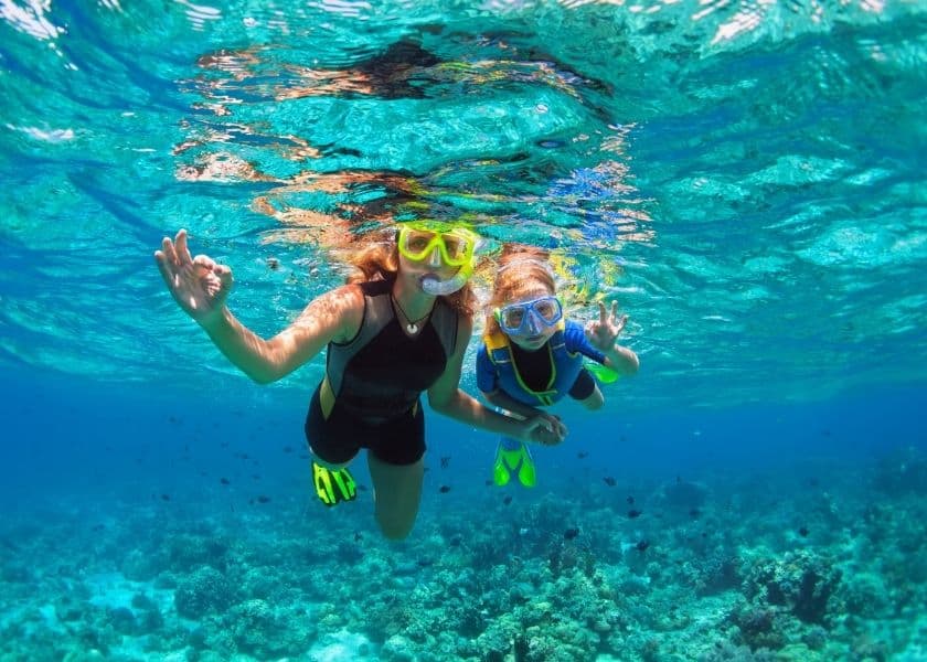 A woman and a child are snorkeling underwater, waving and smiling amidst vibrant blue waters and coral. A woman and a child are snorkeling underwater, waving and smiling amidst vibrant blue waters and coral.