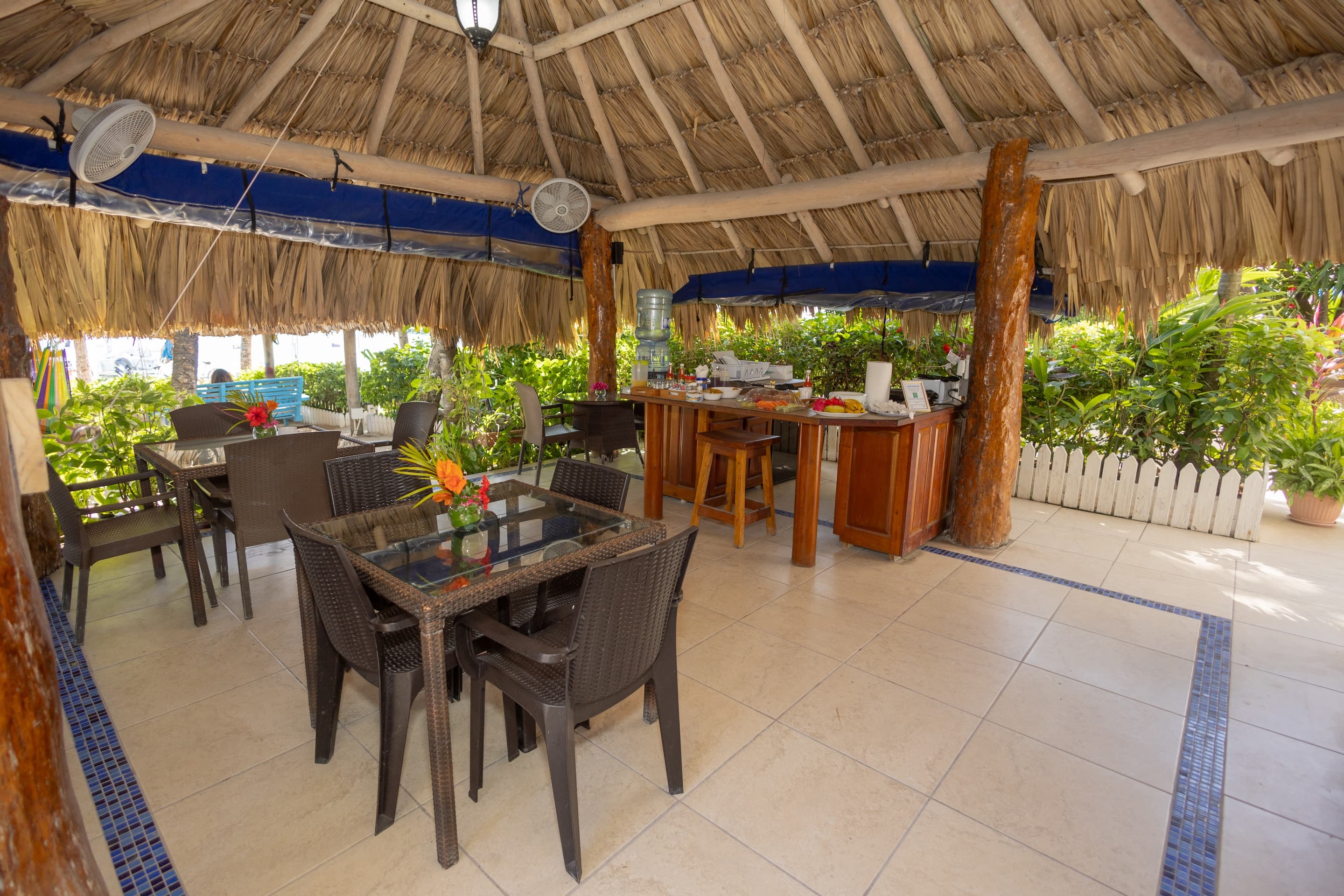 An open-air dining area with a thatched roof, tables and chairs, and a bar area, surrounded by lush greenery.