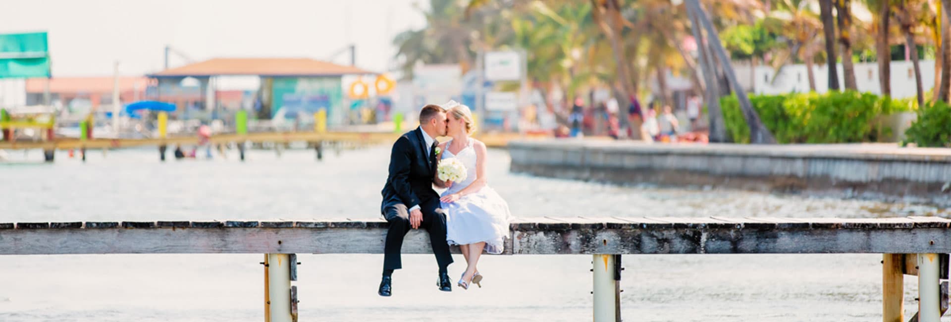 A newlywed couple kisses while sitting on a dock overlooking the water.