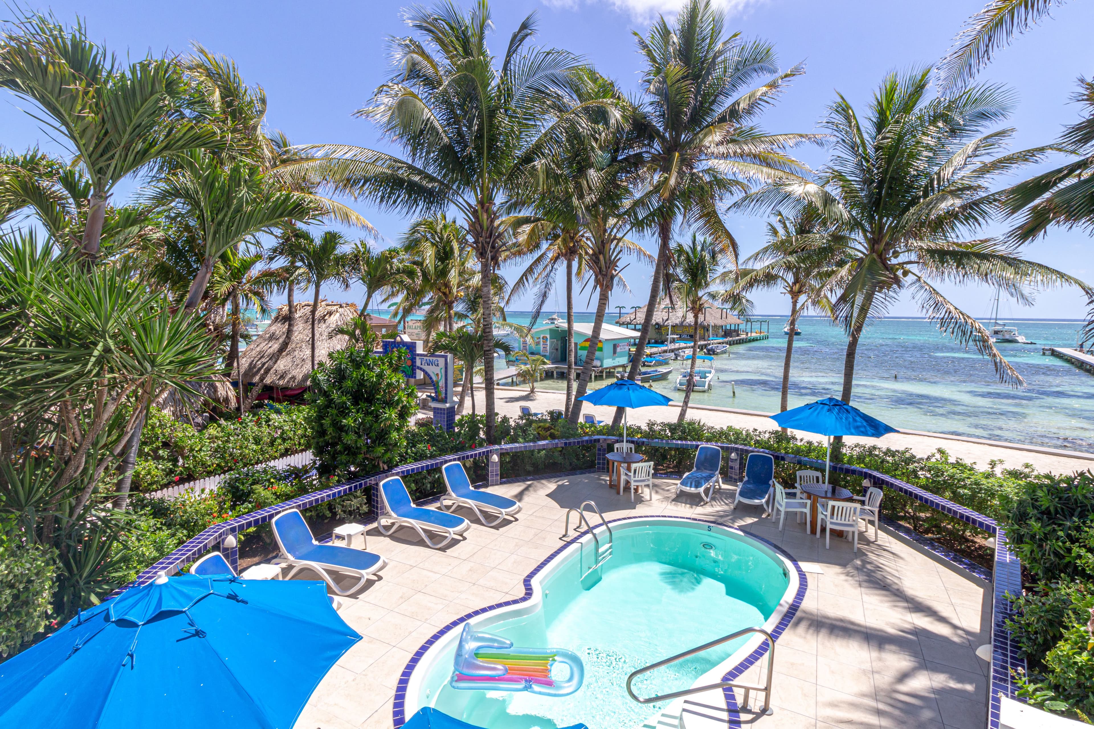 A pool surrounded by palm trees and blue umbrellas, with a view of the ocean and docked boats in the background.