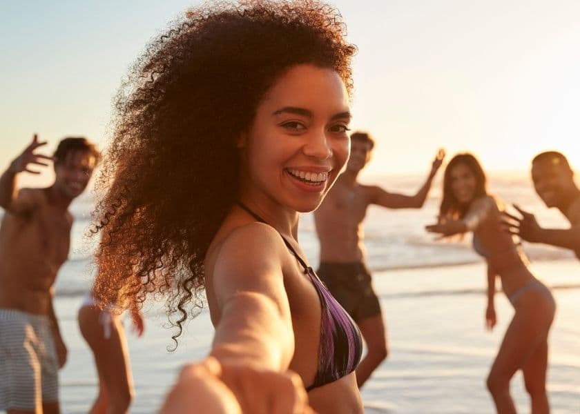 A smiling woman in a bikini reaches out towards the camera with friends playfully posing behind her on a beach at sunset. A smiling woman in a bikini reaches out towards the camera with friends playfully posing behind her on a beach at sunset.