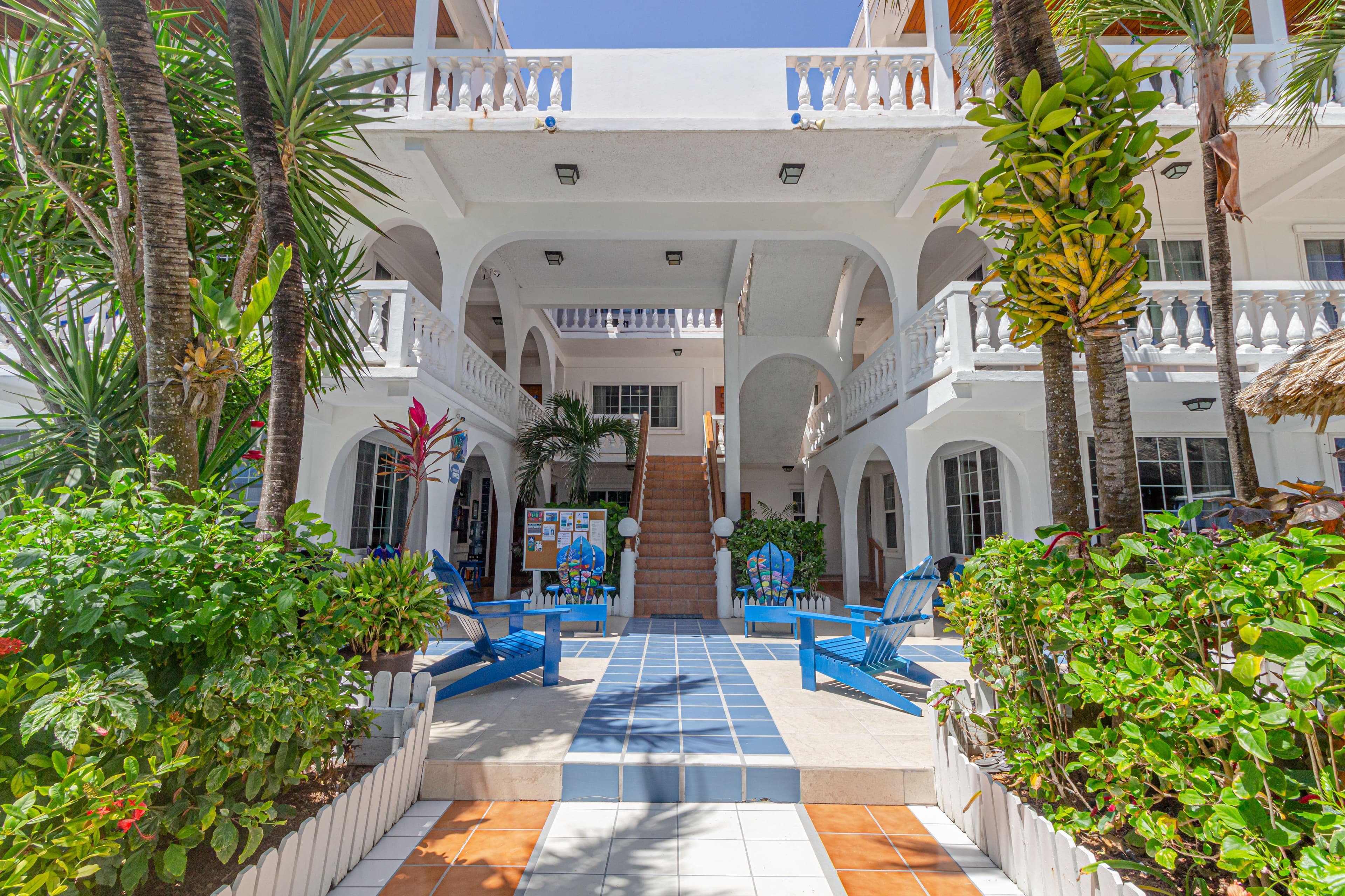 A vibrant courtyard featuring blue chairs, tropical plants, and white architecture under a clear blue sky.