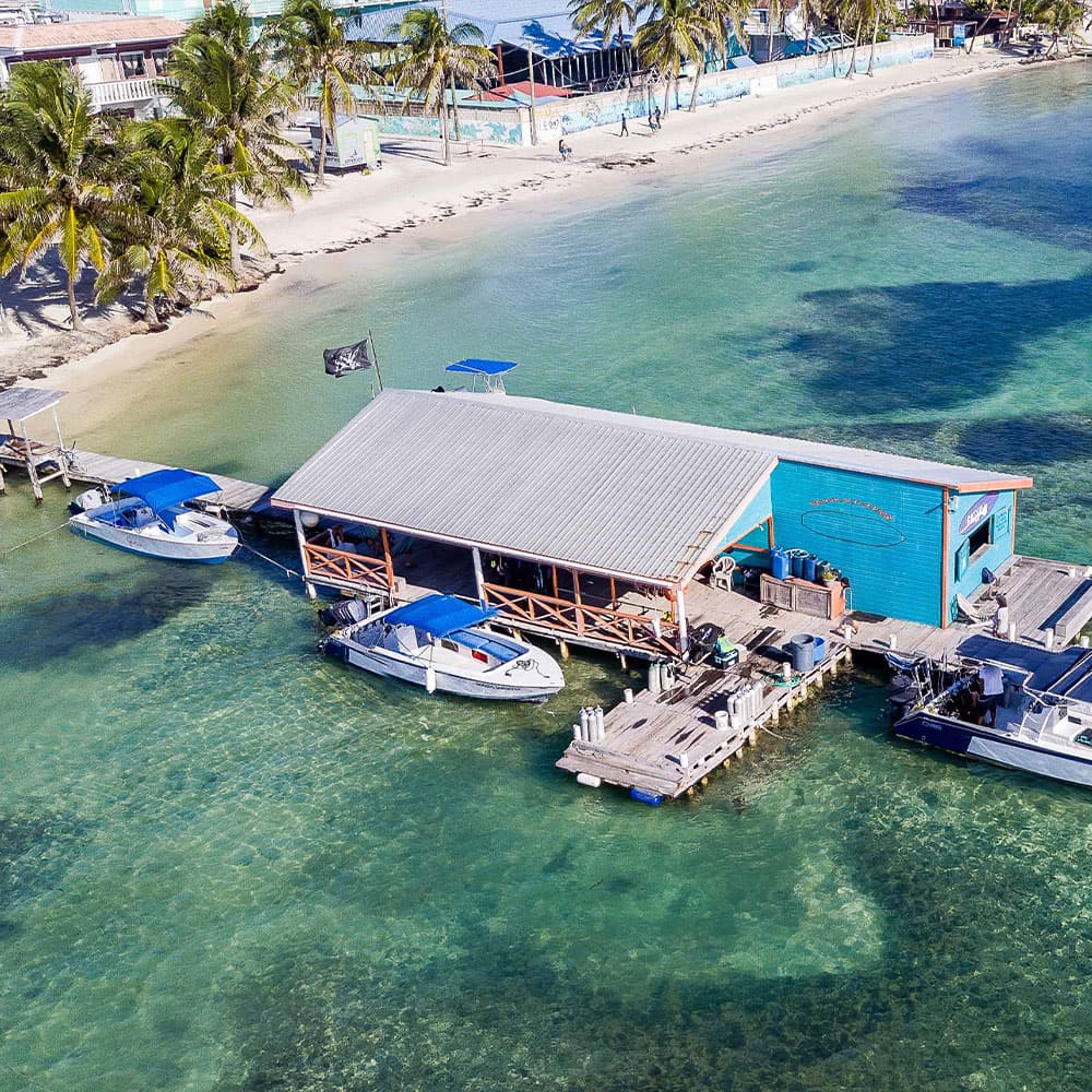 Aerial view of a colorful dockside building with two boats moored nearby, surrounded by clear turquoise waters and palm trees.