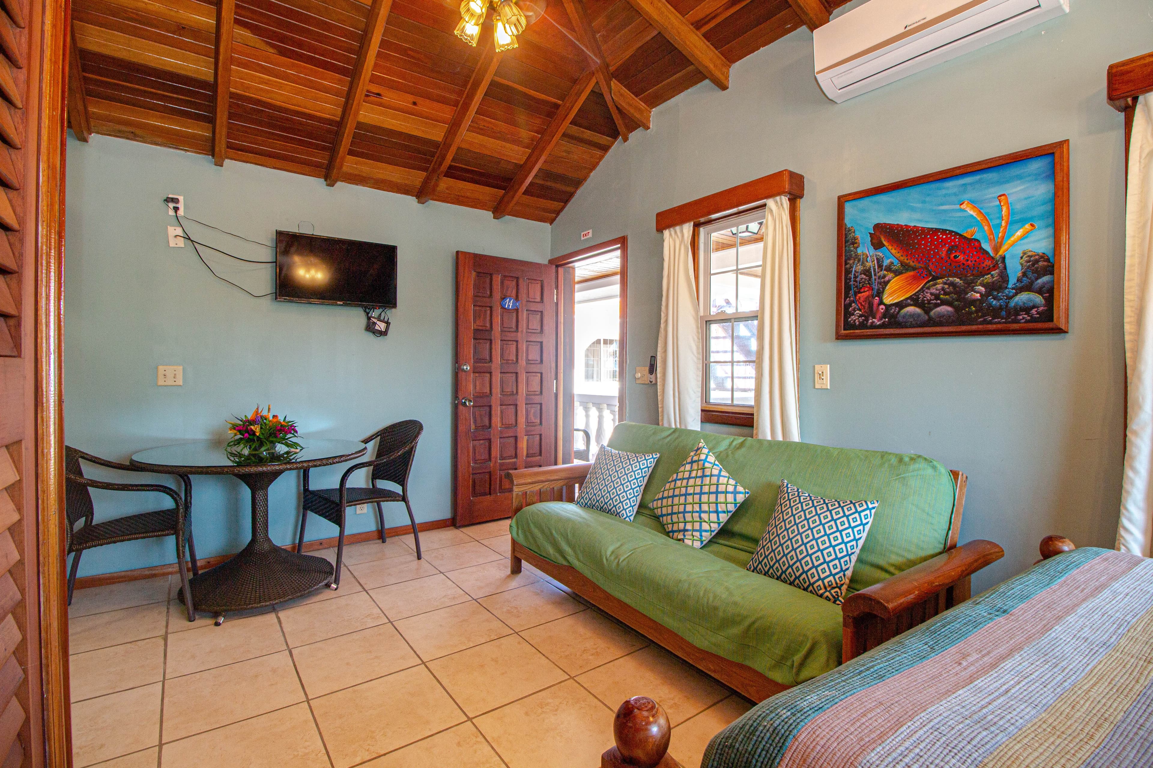 A room with a high wooden ceiling, a green futon, a round table with chairs, a mounted TV, and a painting on the wall.