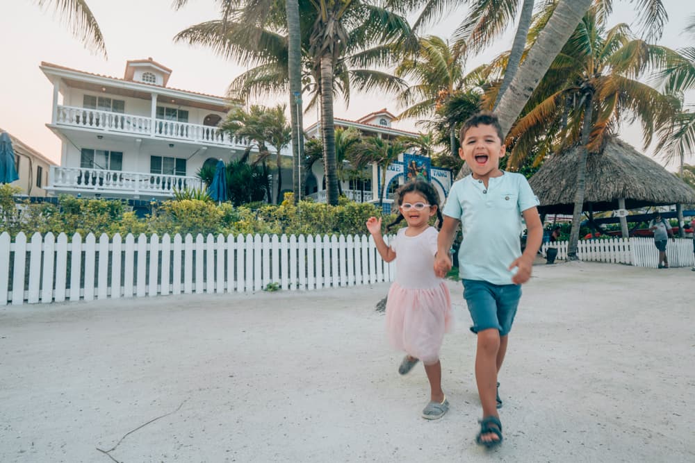 Two children joyfully running along a sandy path in front of a white picket fence and tropical buildings.
