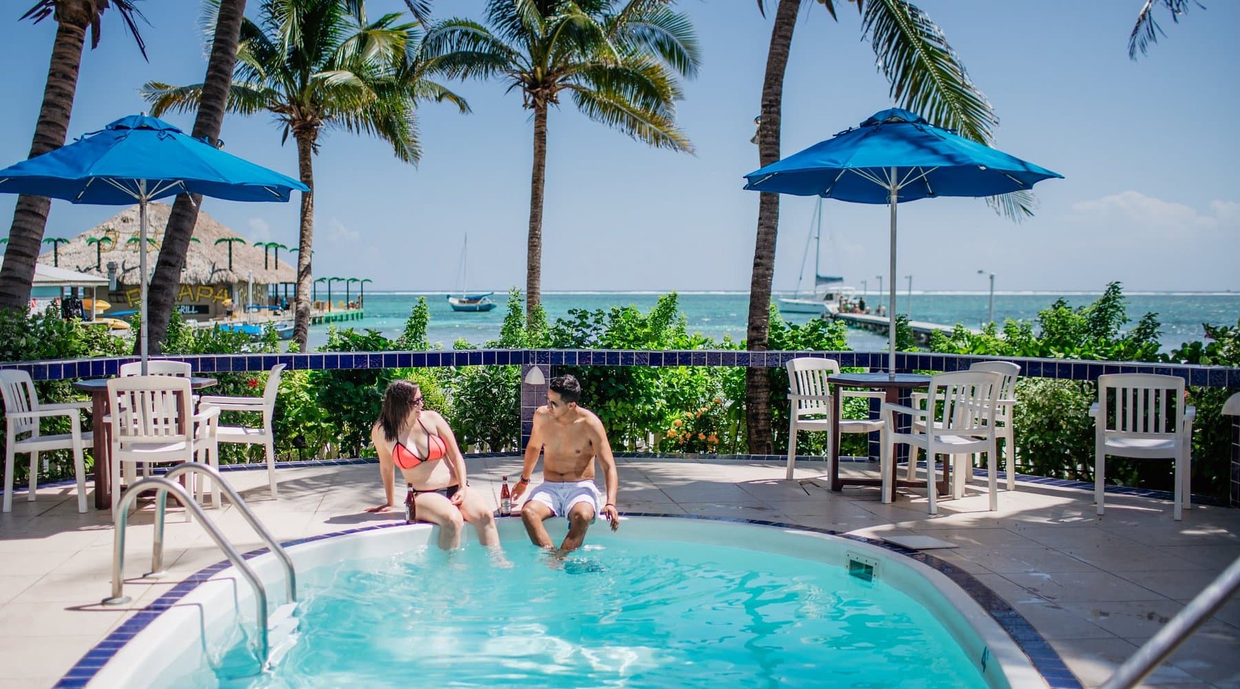 A couple sits by a pool with drinks, surrounded by palm trees and a scenic ocean view.