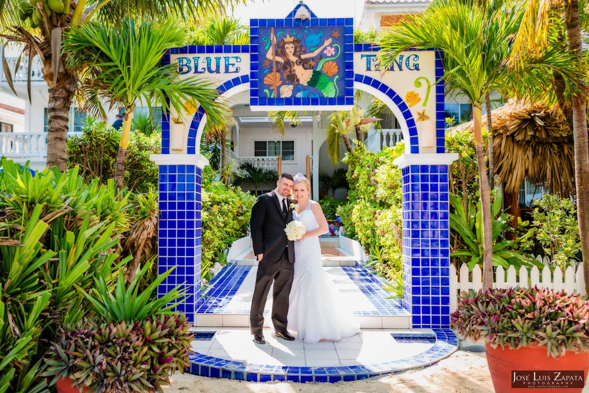 A newlywed couple stands under a colorful tiled archway surrounded by lush greenery.