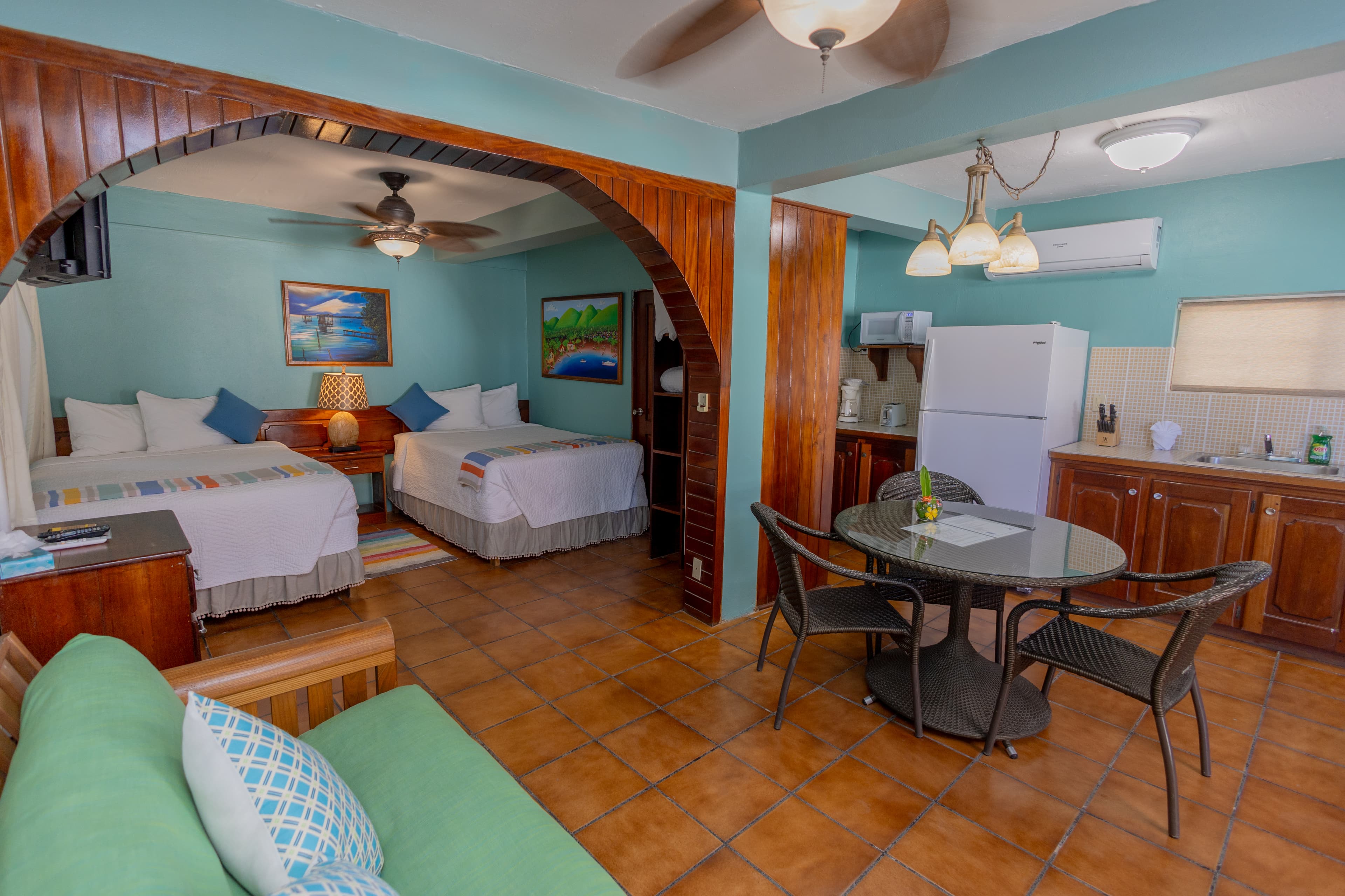 A cozy hotel room featuring two beds, a small dining area, and a kitchenette with light blue walls and wooden accents.