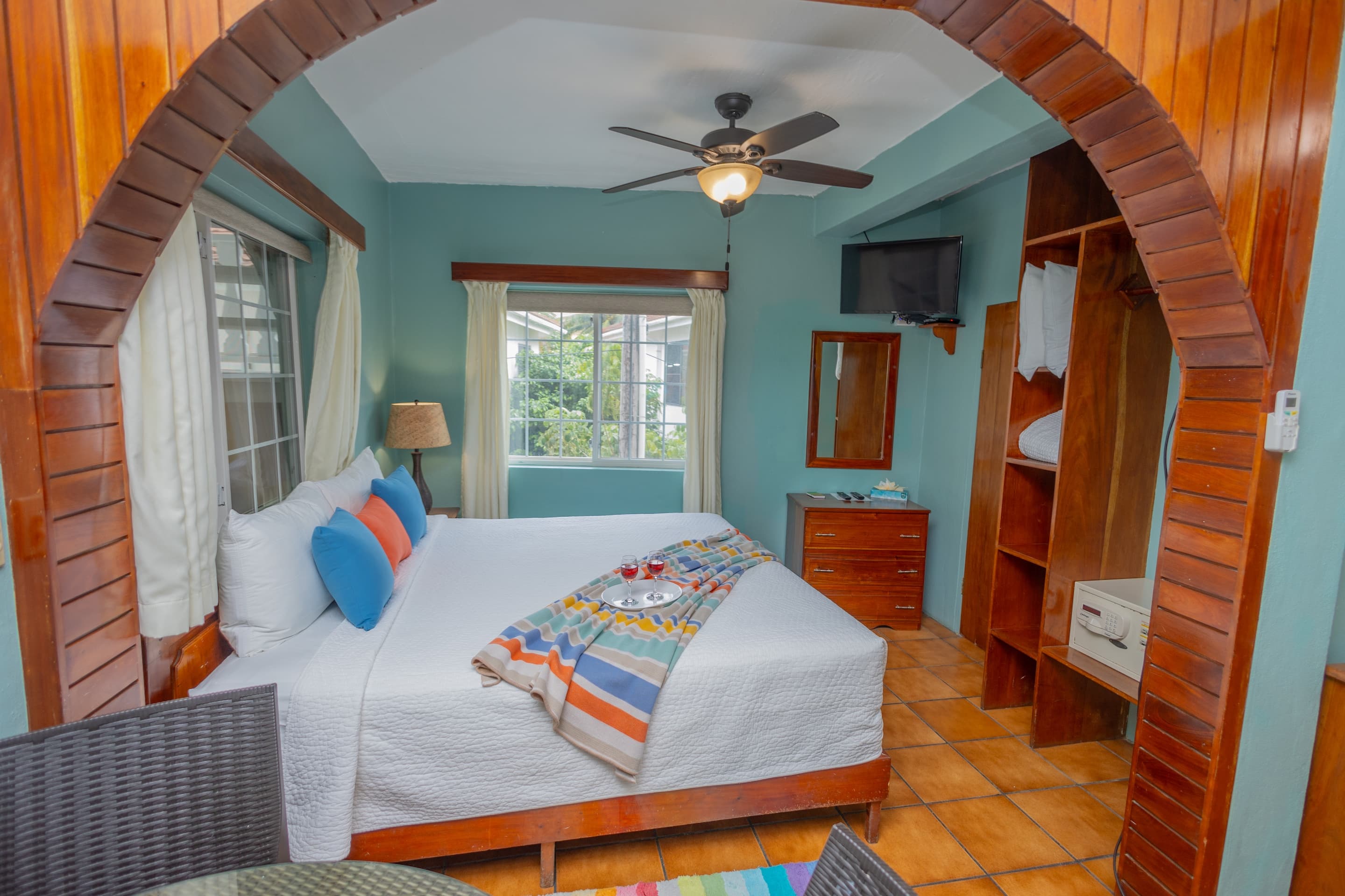 A bedroom with teal walls, tiled floors, a king bed with colorful pillows and runner, a ceiling fan, and a wooden archway leading to another area.