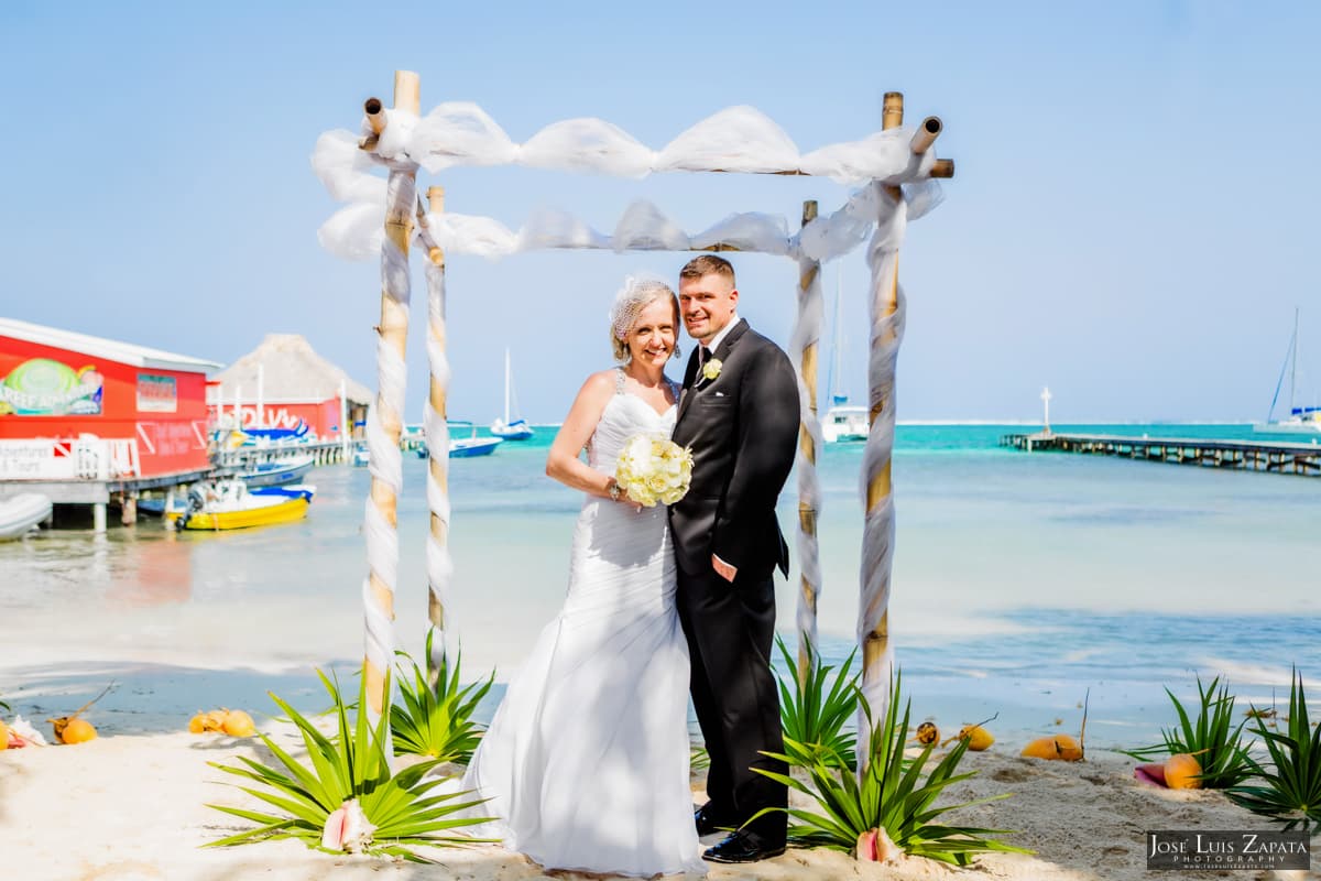 A happy couple poses under a decorated wedding arch on the beach with turquoise waters in the background.
