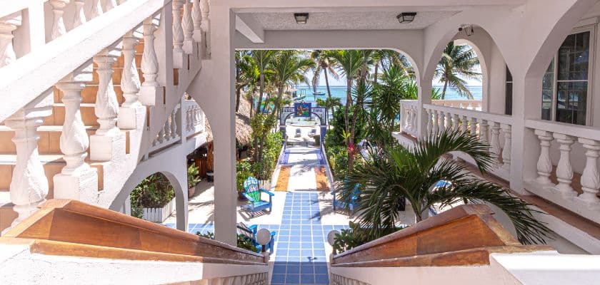 A view of a tropical courtyard with palm trees and a pathway leading to the beach, framed by white stairs and arches.