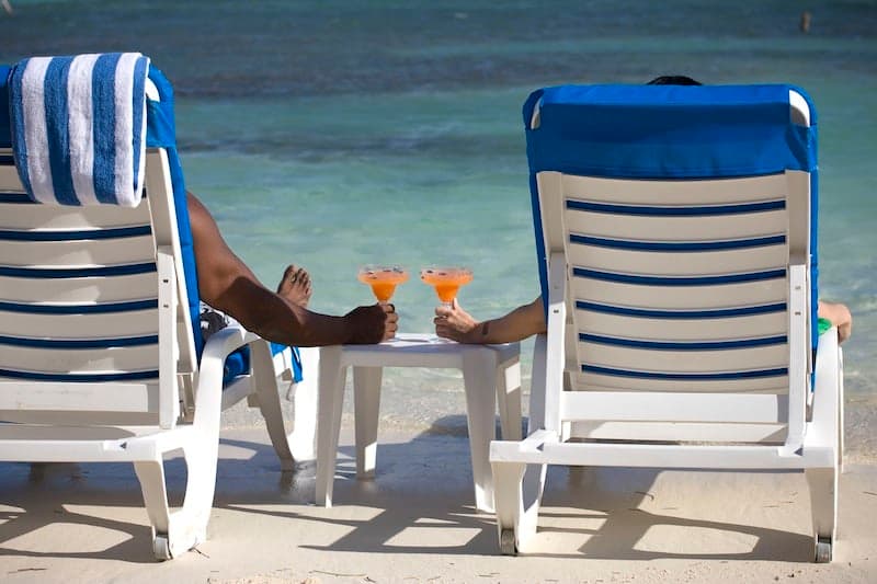 Two people relax in beach chairs holding cocktails, with a calm ocean in the background.