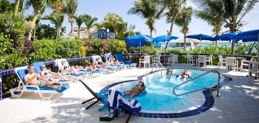 A sunny poolside scene with several people lounging on chairs surrounded by palm trees and blue umbrellas.