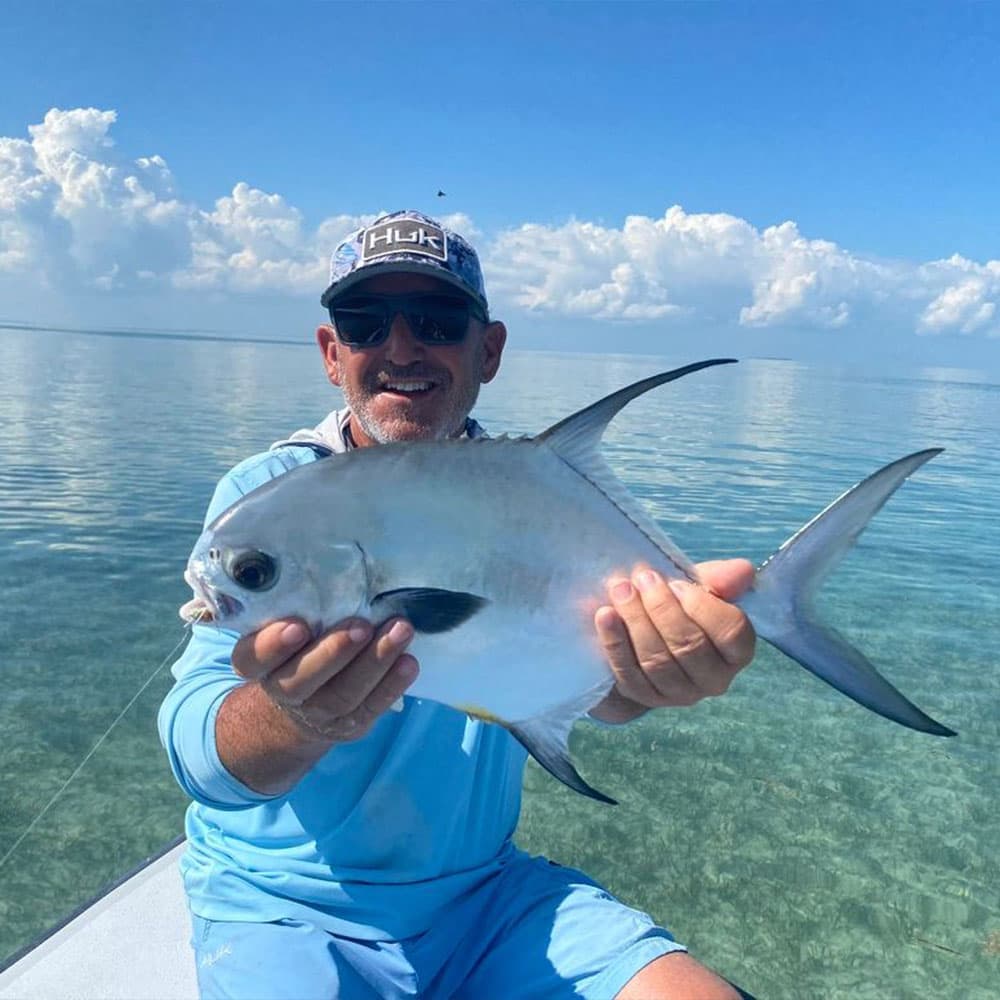 A smiling man in sunglasses holds a large fish on a boat in clear water under a blue sky.