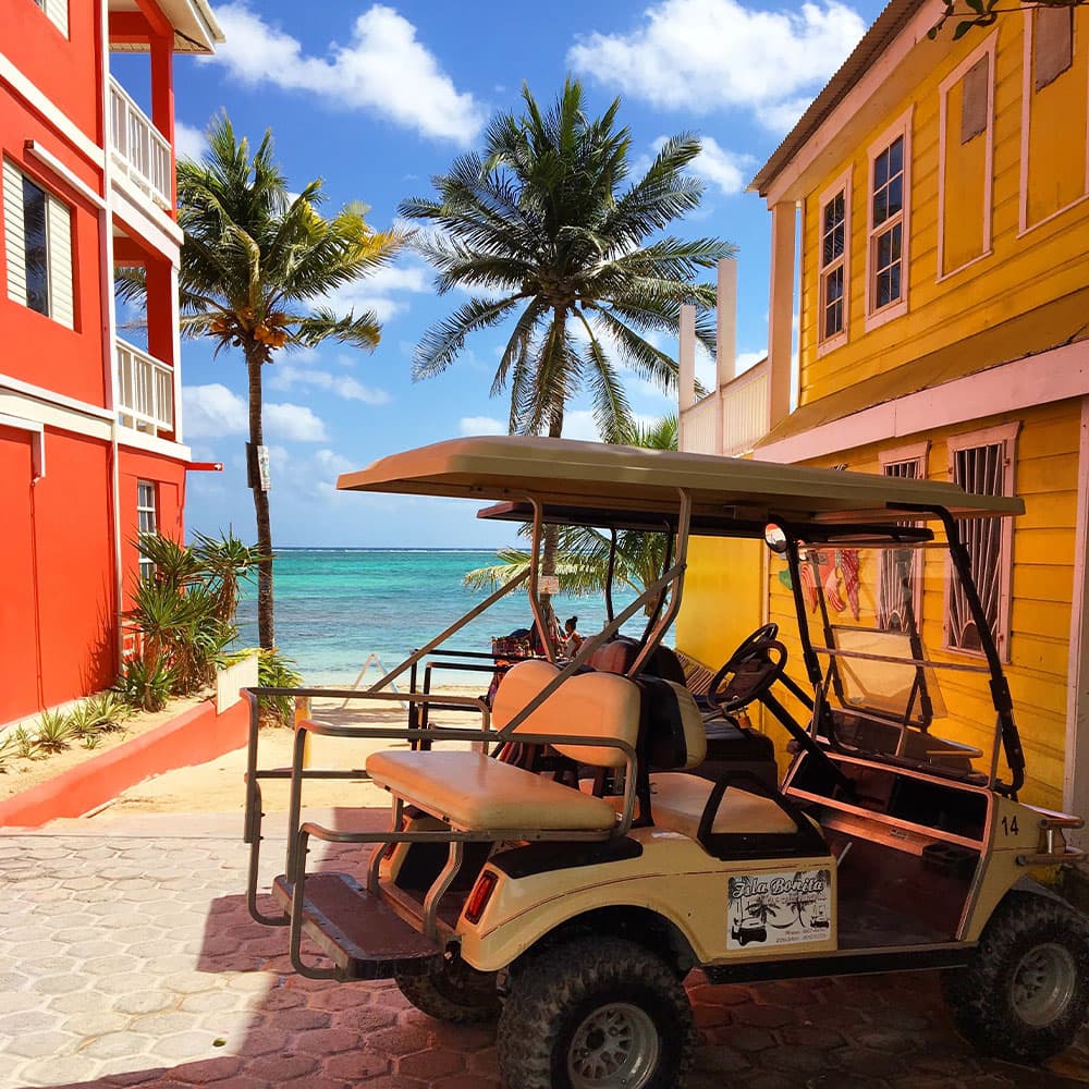 A golf cart is parked beside colorful buildings with palm trees and a beach in the background.