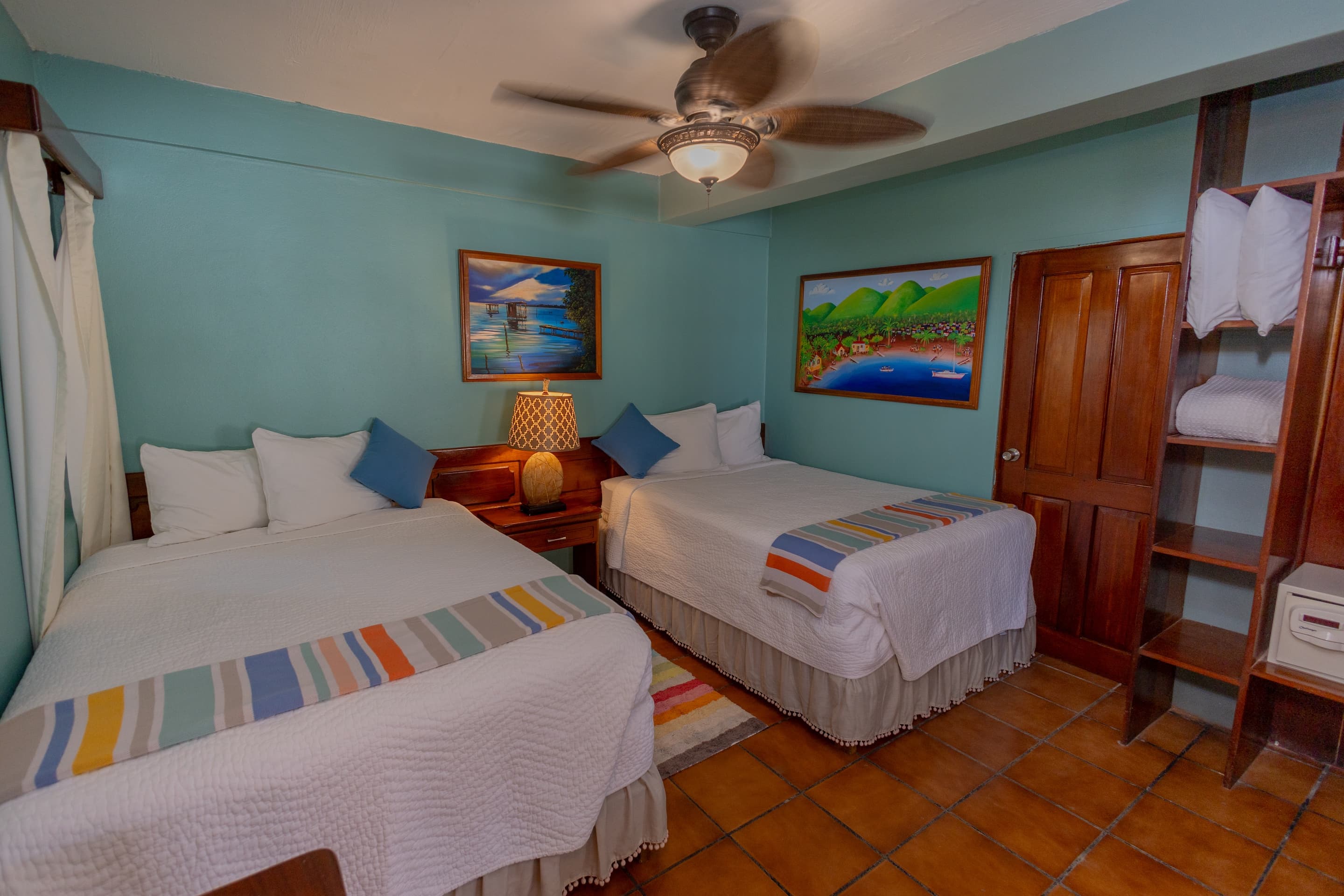 A bedroom with teal walls, tiled floors, two full beds with colorful accents, a ceiling fan, and framed artwork.