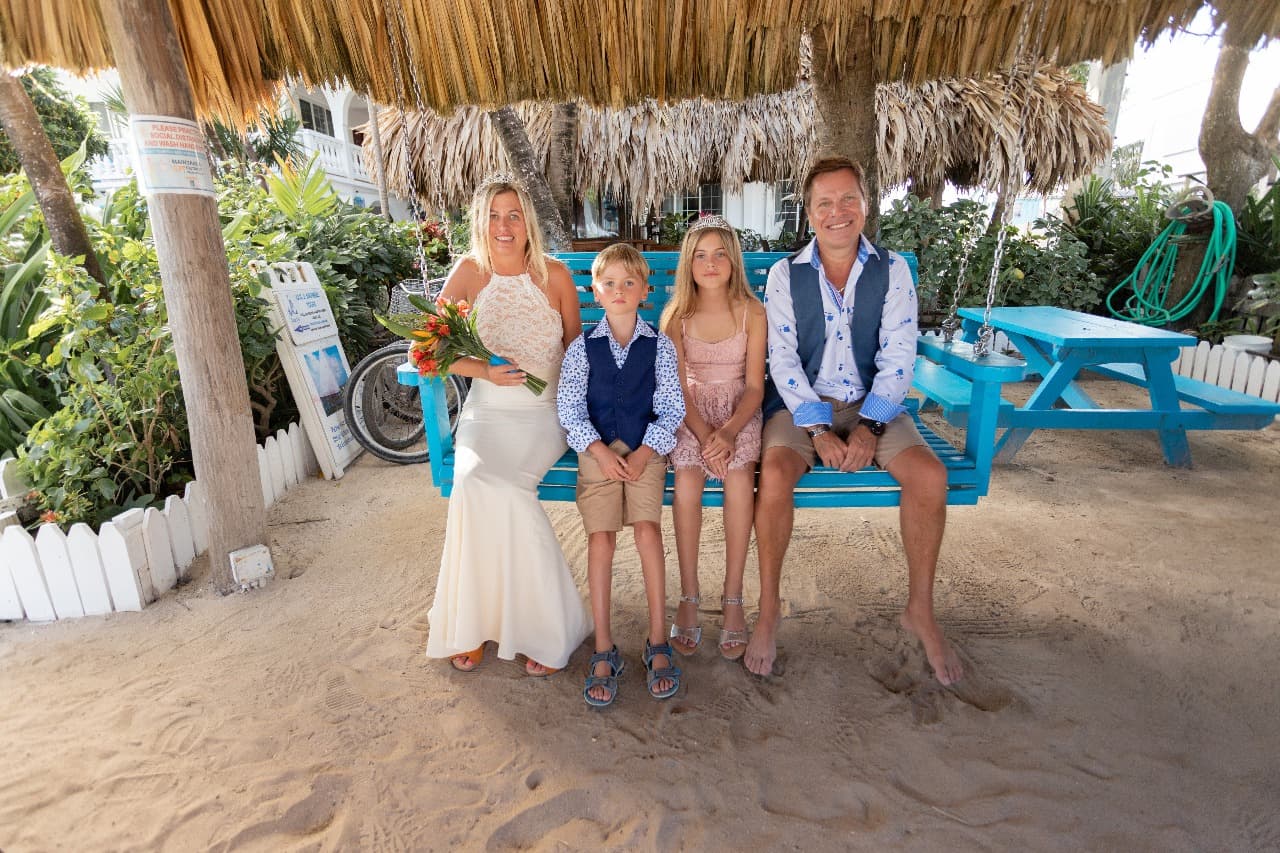 A family of four poses together on a swing under a thatched roof with greenery in the background.