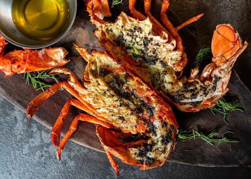 A cooked lobster topped with seasoning, accompanied by a small bowl of olive oil on a wooden platter. A cooked lobster topped with seasoning, accompanied by a small bowl of olive oil on a wooden platter.