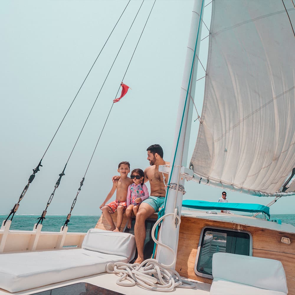 A man sits on a sailboat with two smiling children against a backdrop of calm water and a clear sky.