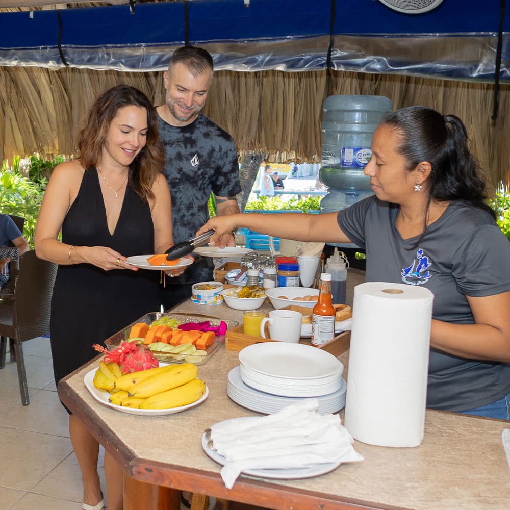 A woman in a black dress is receiving food on a plate from a server while a man smiles in the background at a buffet table with various food items.