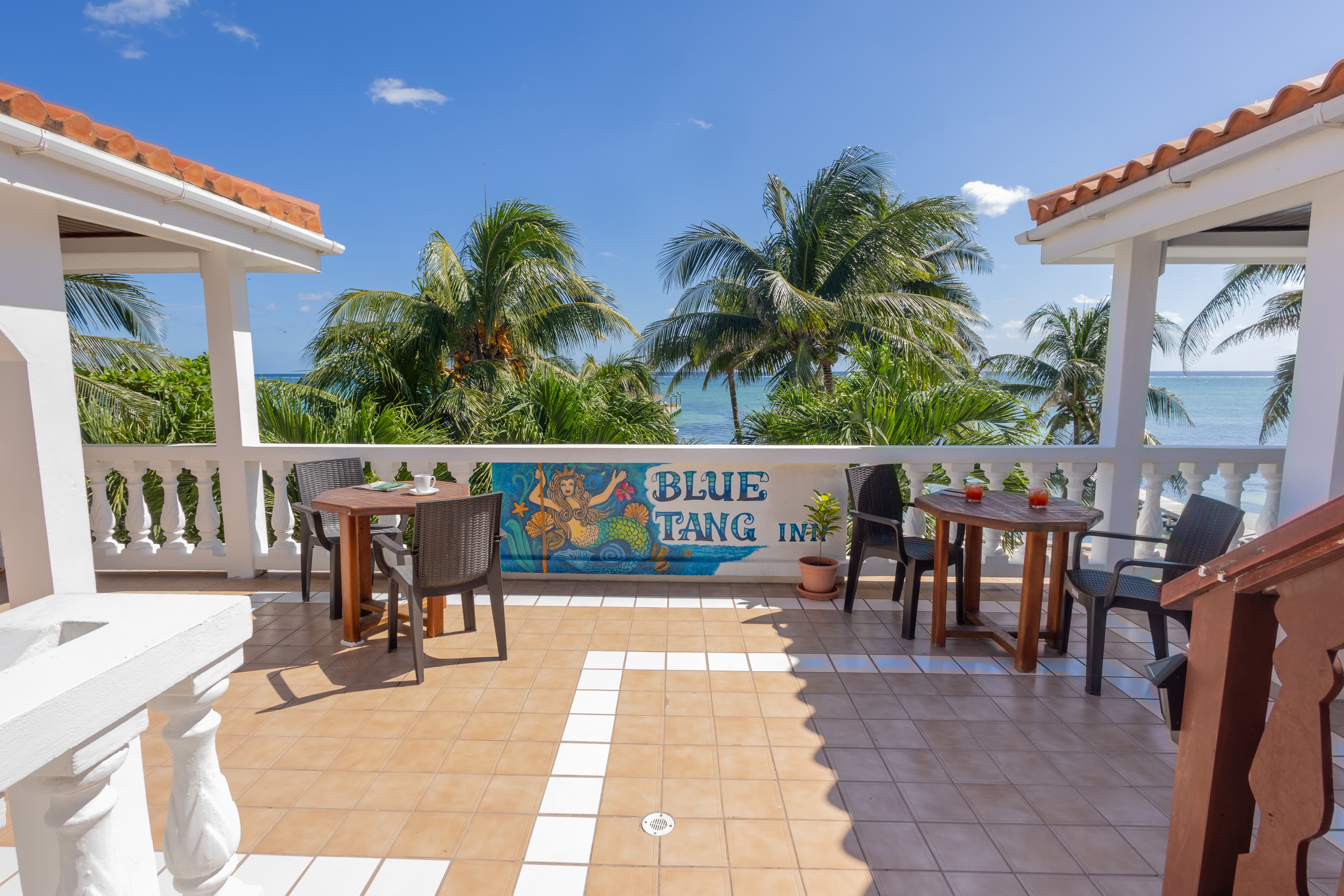 A sunlit balcony with palm trees and a view of the ocean features two tables and a mural reading "Blue Tang Inn."