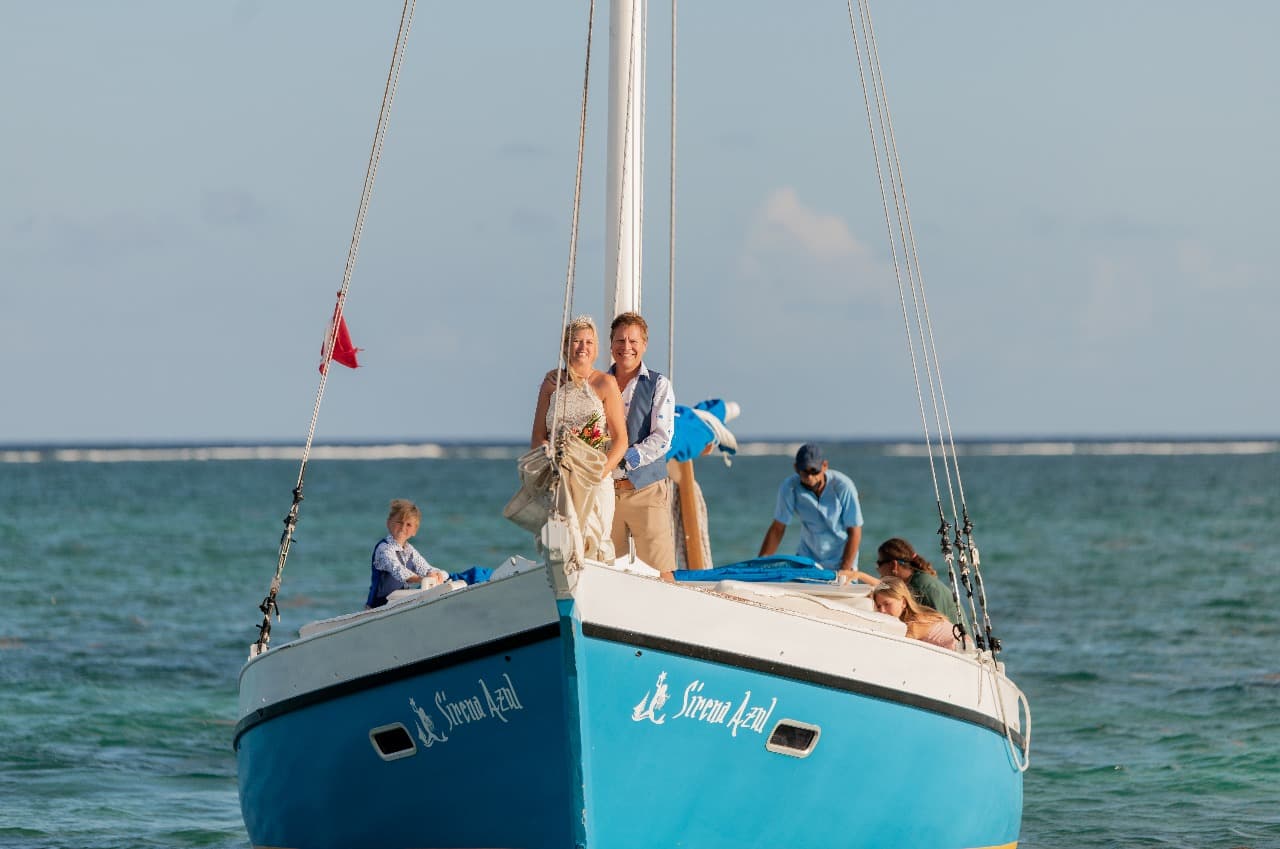 A couple stands on the bow of a sailboat, surrounded by guests, on a sunny ocean.