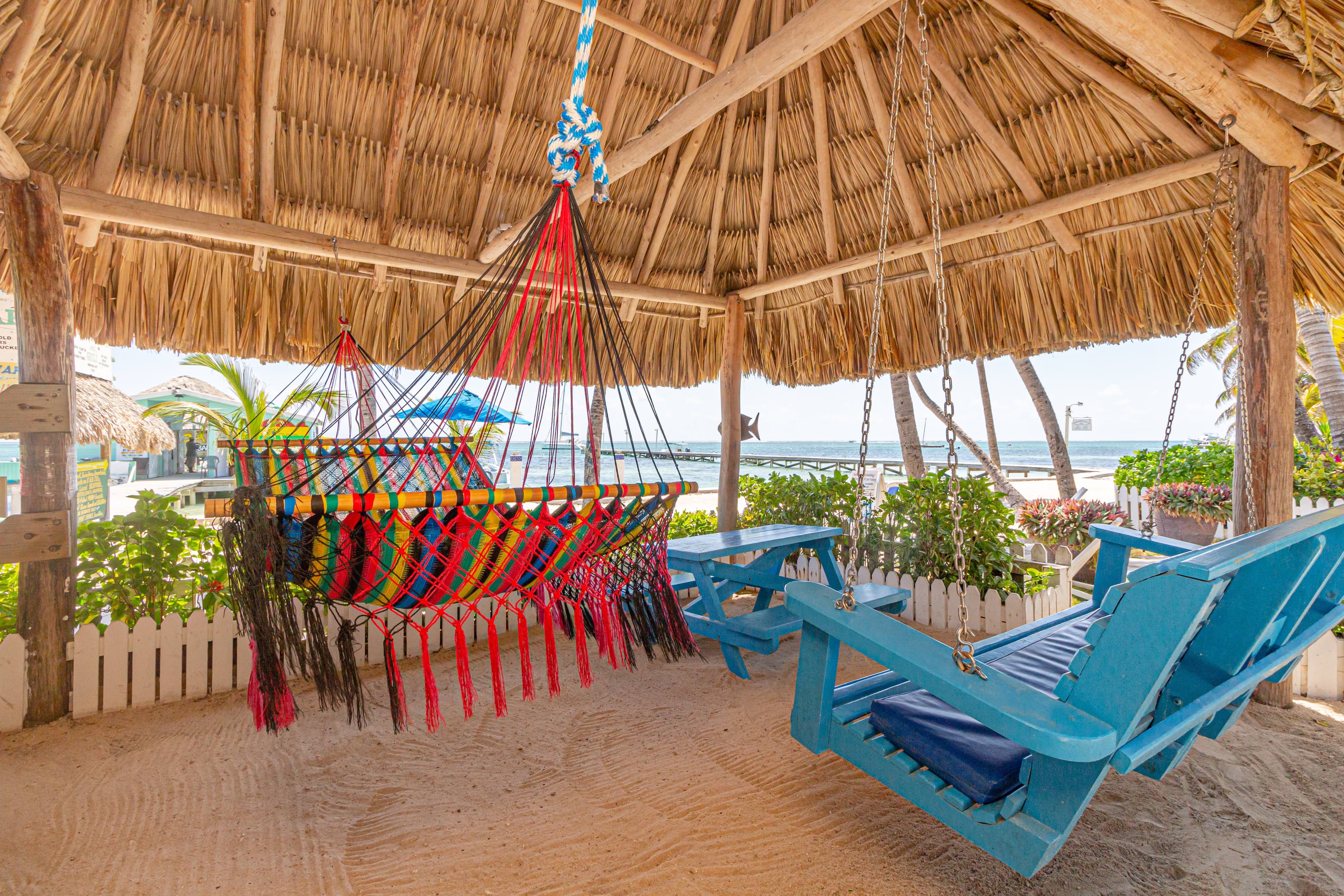 A colorful hammock hangs under a thatched roof near blue wooden chairs and a sandy beach.