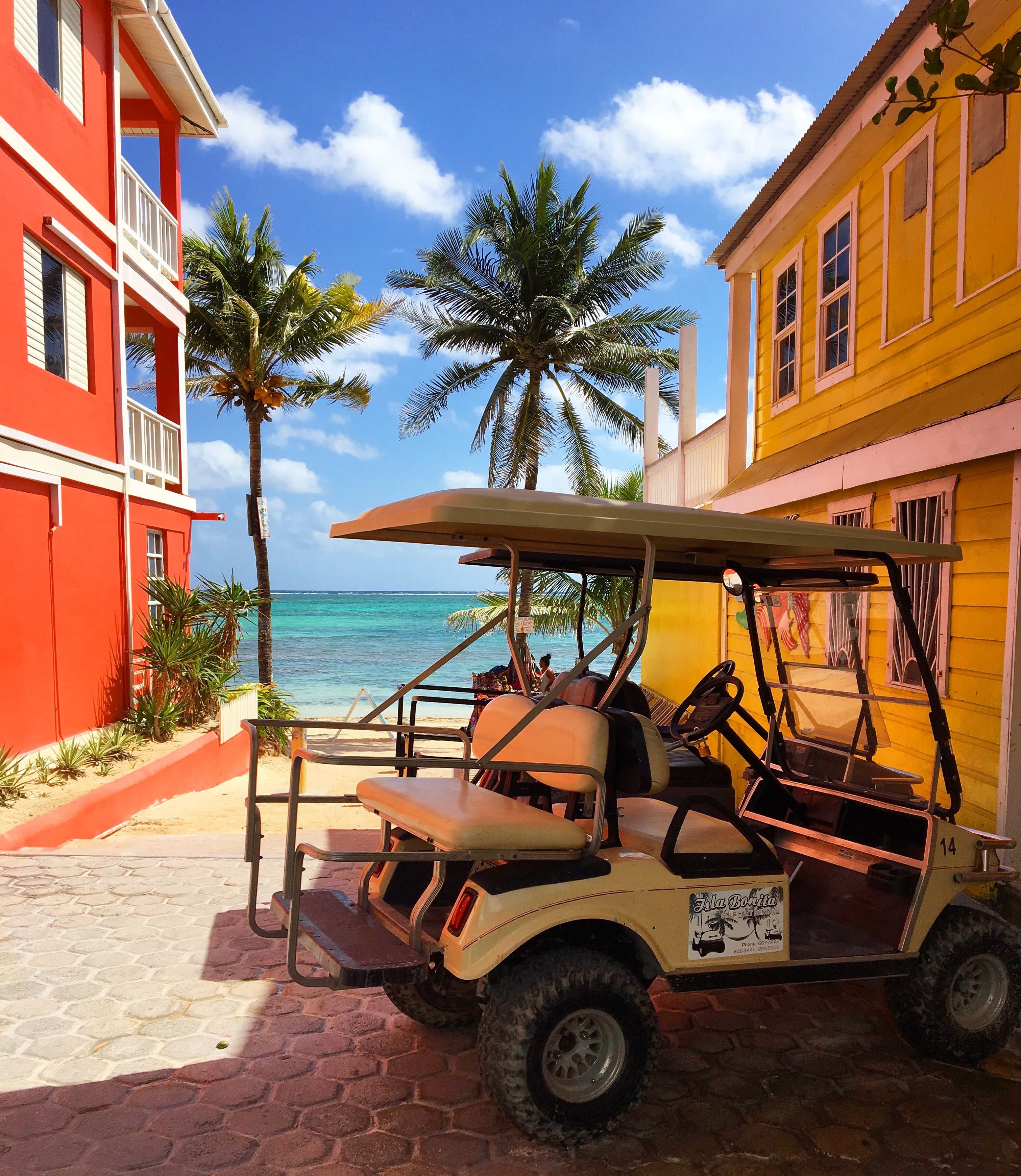 A golf cart is parked between colorful buildings near a tropical beach.