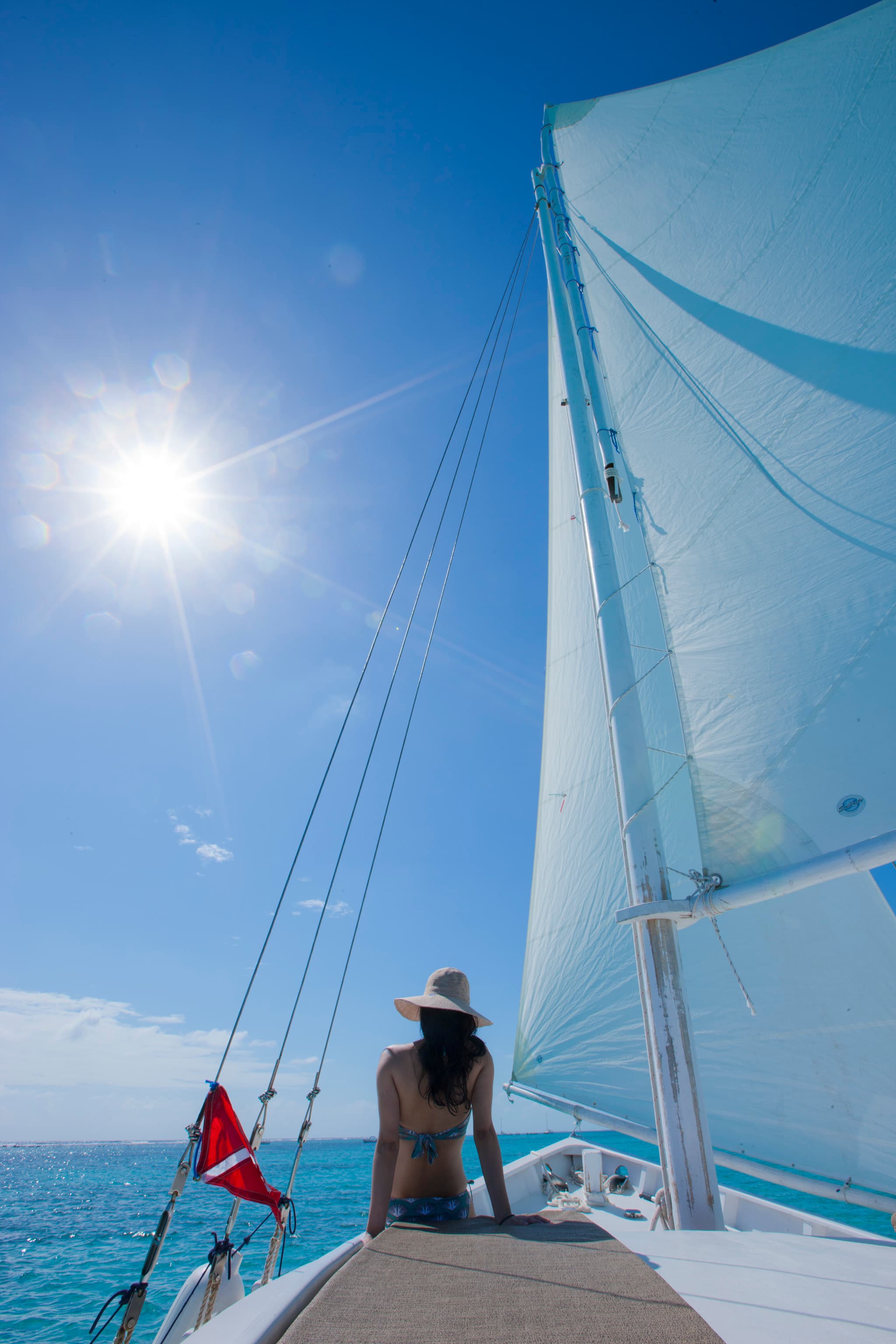 A person in a hat sits on the edge of a sailboat, gazing at the sunlit ocean.