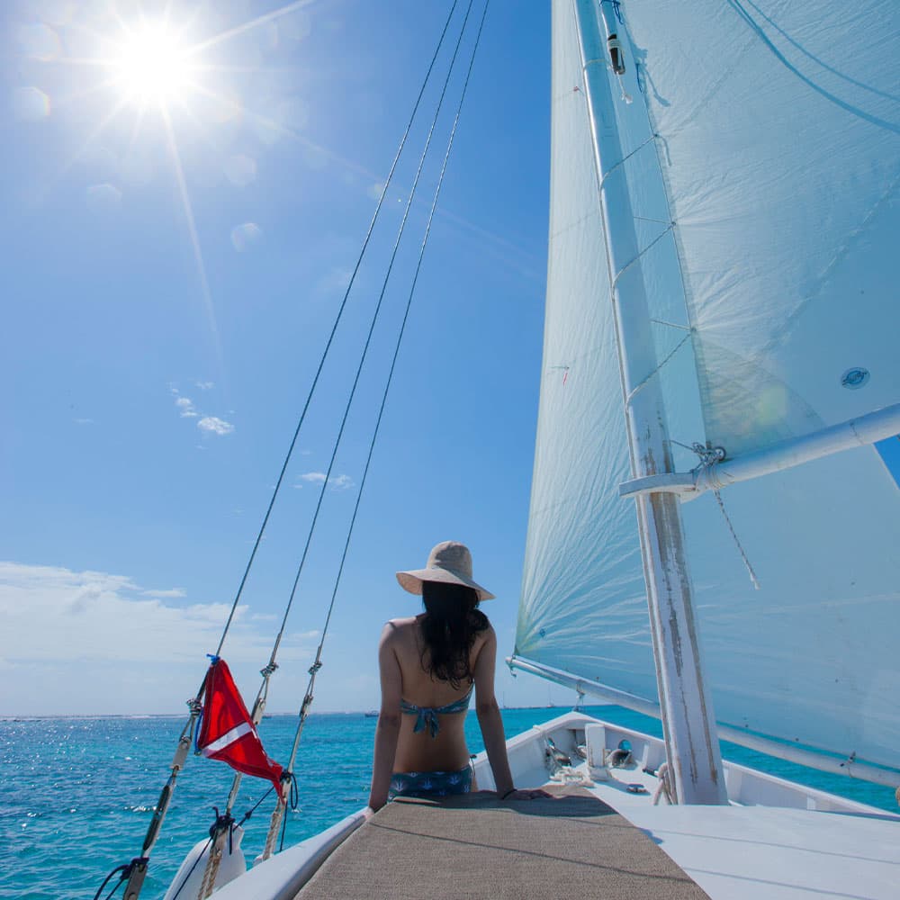A person sits on a sailboat, gazing at the bright blue sea under a sunny sky.