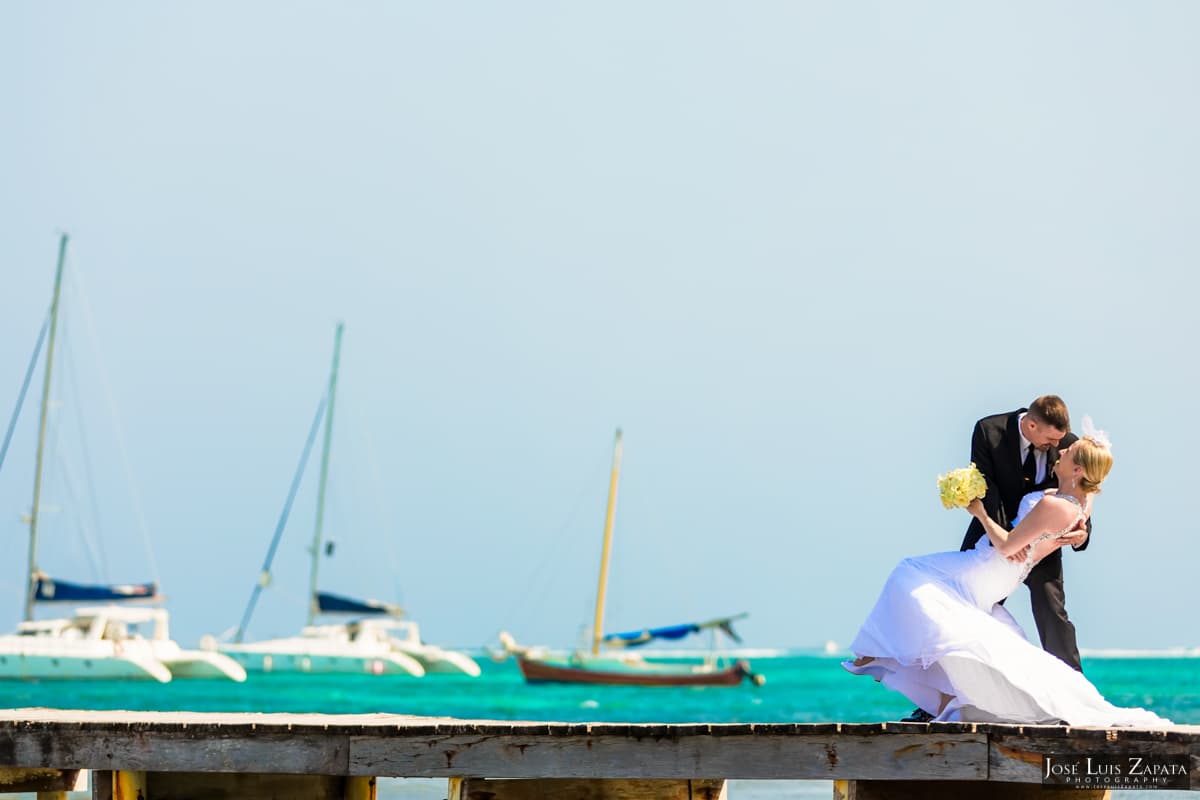 A couple embraces on a wooden pier with sailboats and turquoise water in the background.