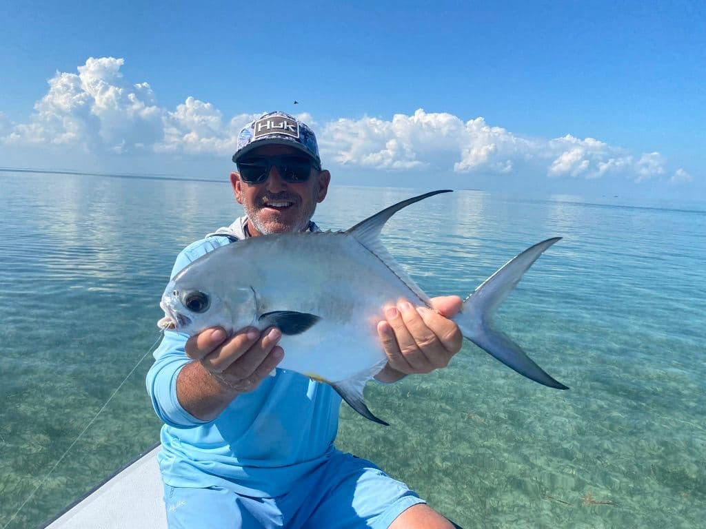 A man in a blue shirt holds a fish while standing on a boat in calm, clear water under a blue sky.