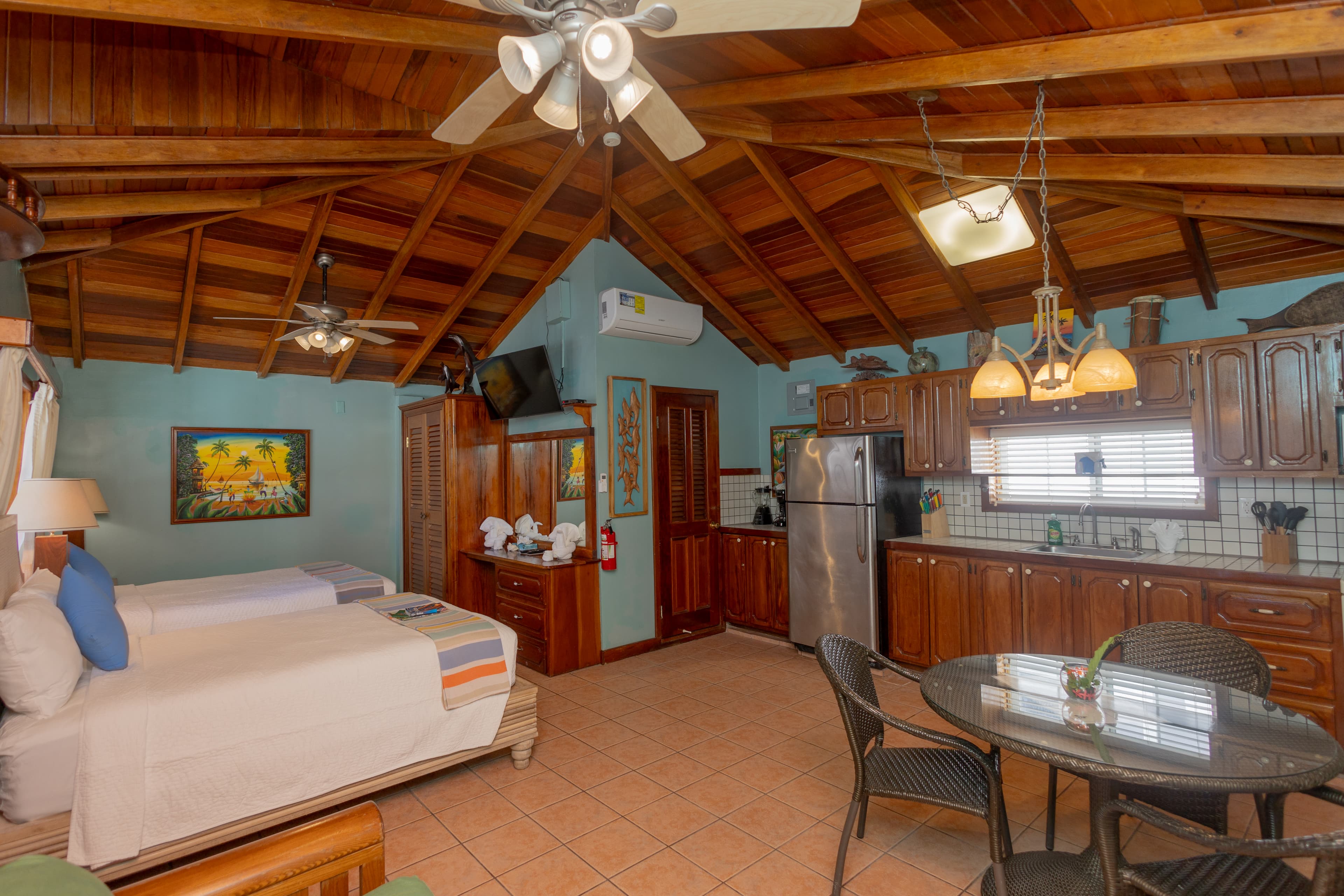 A cozy room featuring two beds, kitchen area, dining table, and wooden ceiling.