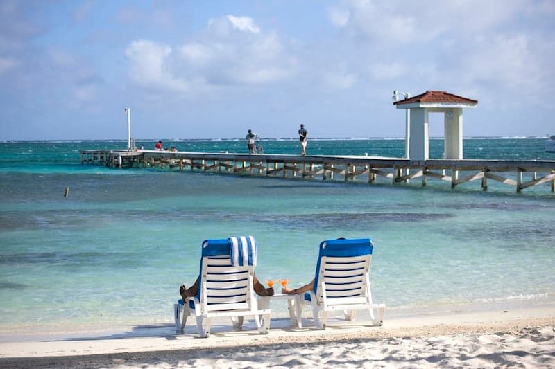 Two beach chairs in front of a pier extending into turquoise waters under a cloudy sky.