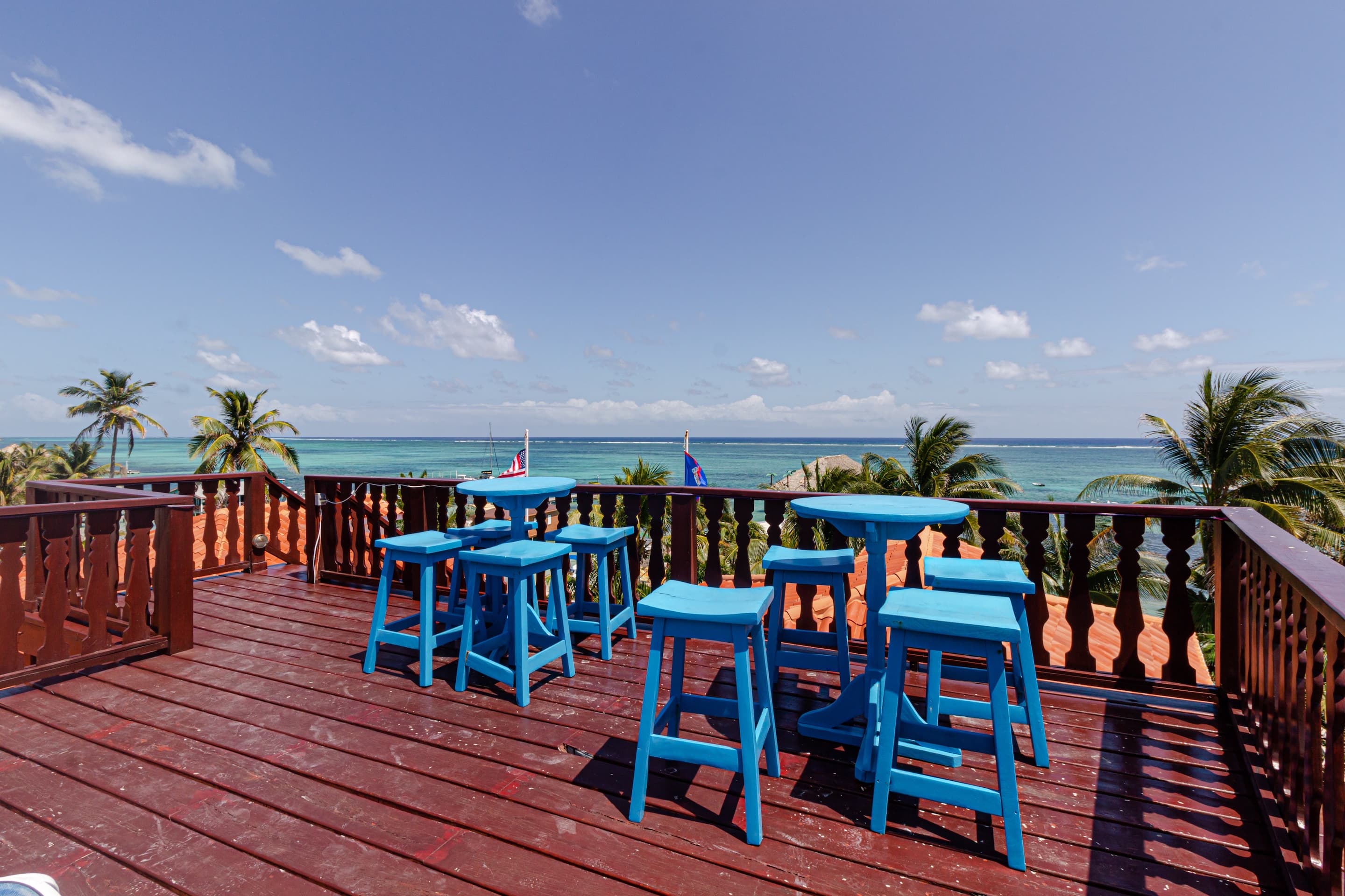 A wooden rooftop deck with blue bar stools and small tables overlooking the ocean and palm trees under a blue sky.