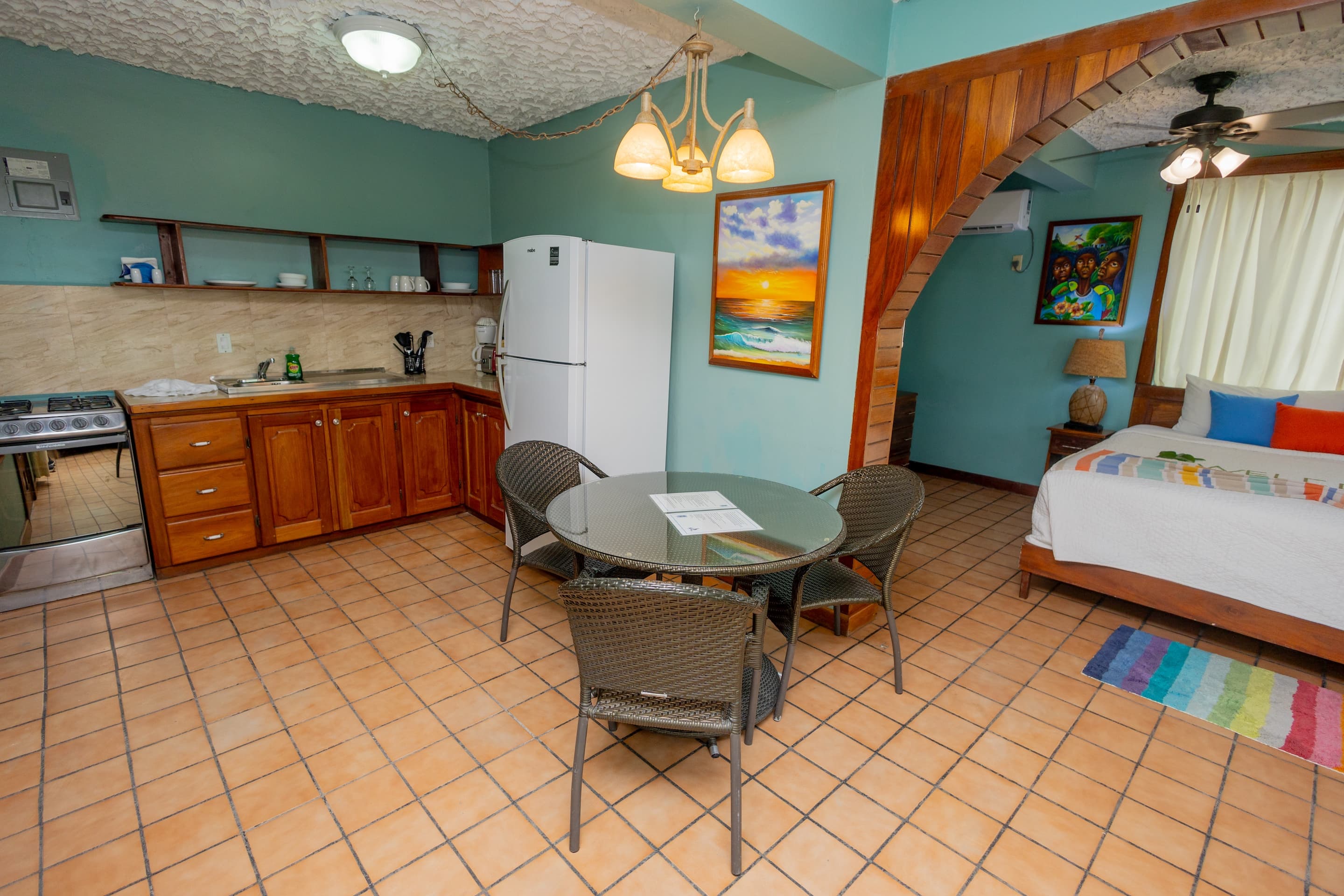 A studio apartment with teal walls, tiled floors, a kitchenette, a dining table with chairs, and a king bed in an adjacent area.
