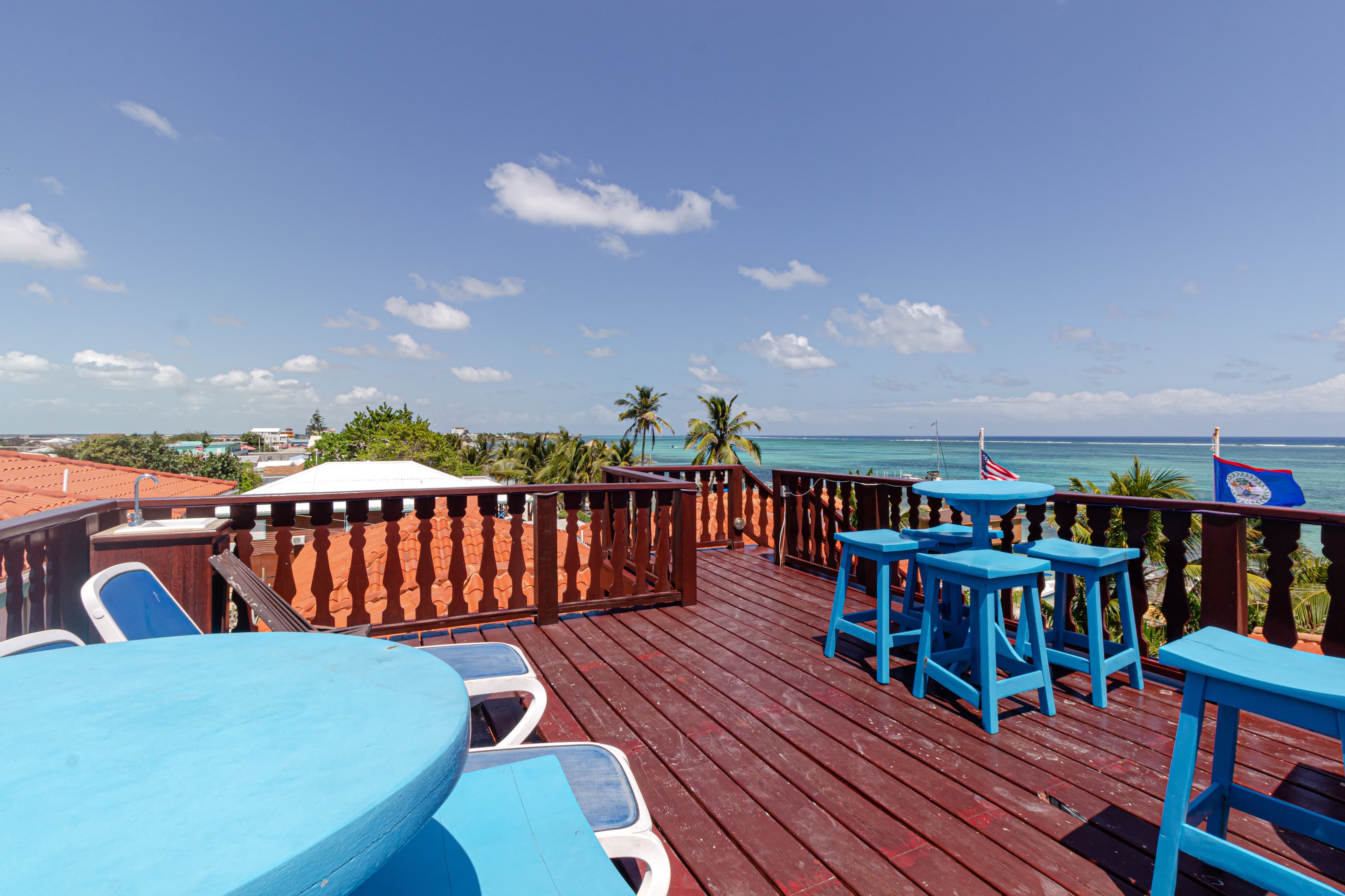 A sunny rooftop terrace with blue tables overlooks the ocean and palm trees.