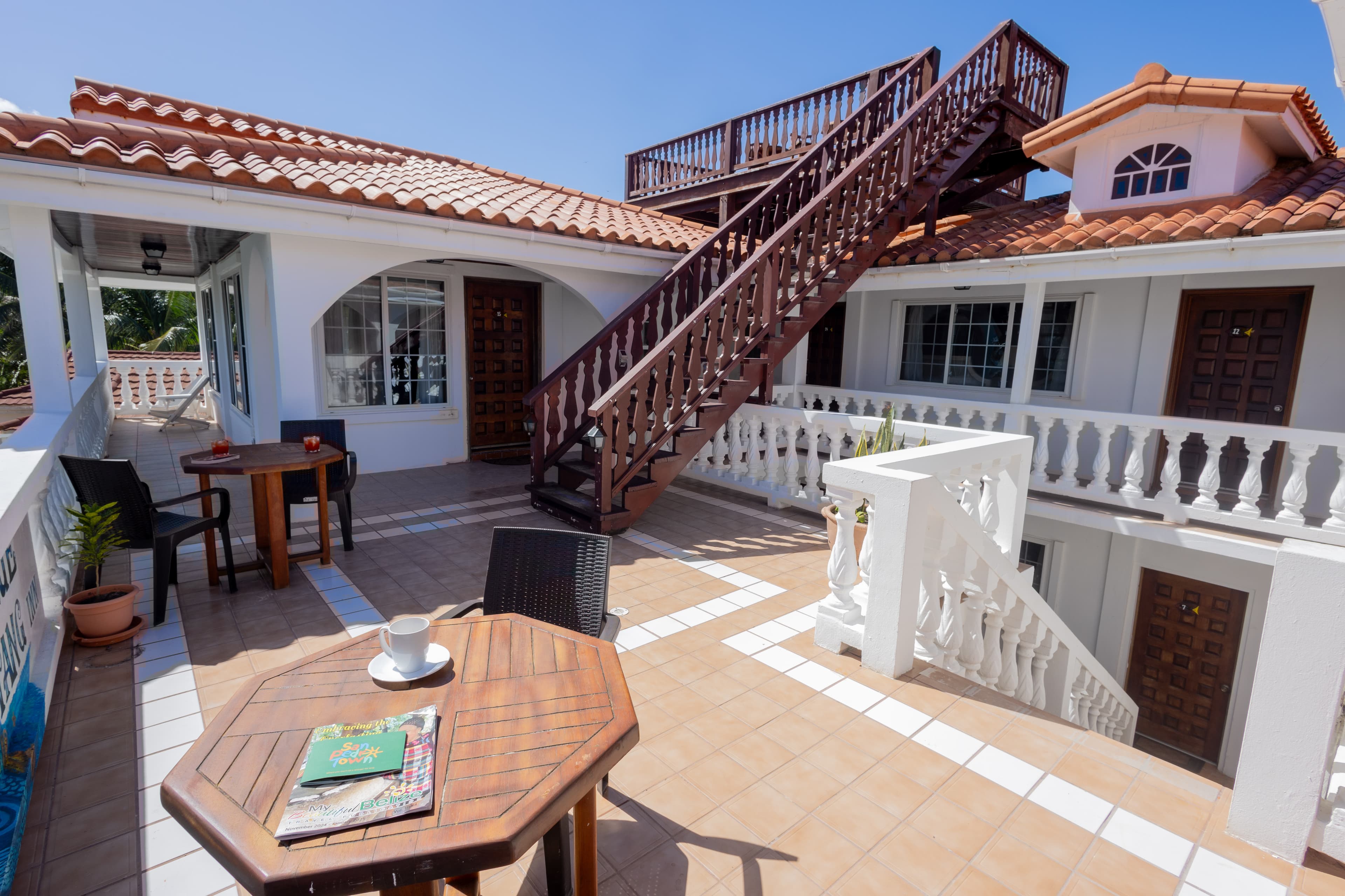 A sunny terrace features a wooden table, a coffee cup, a magazine, and a staircase leading to an upper level.