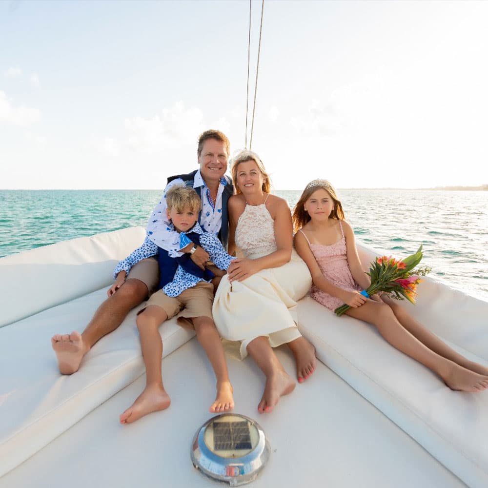 A happy family of four sits on a sailboat, enjoying a sunny day by the water.