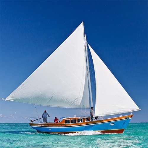A colorful sailboat with three people on board glides across a clear blue ocean under a sunny sky.