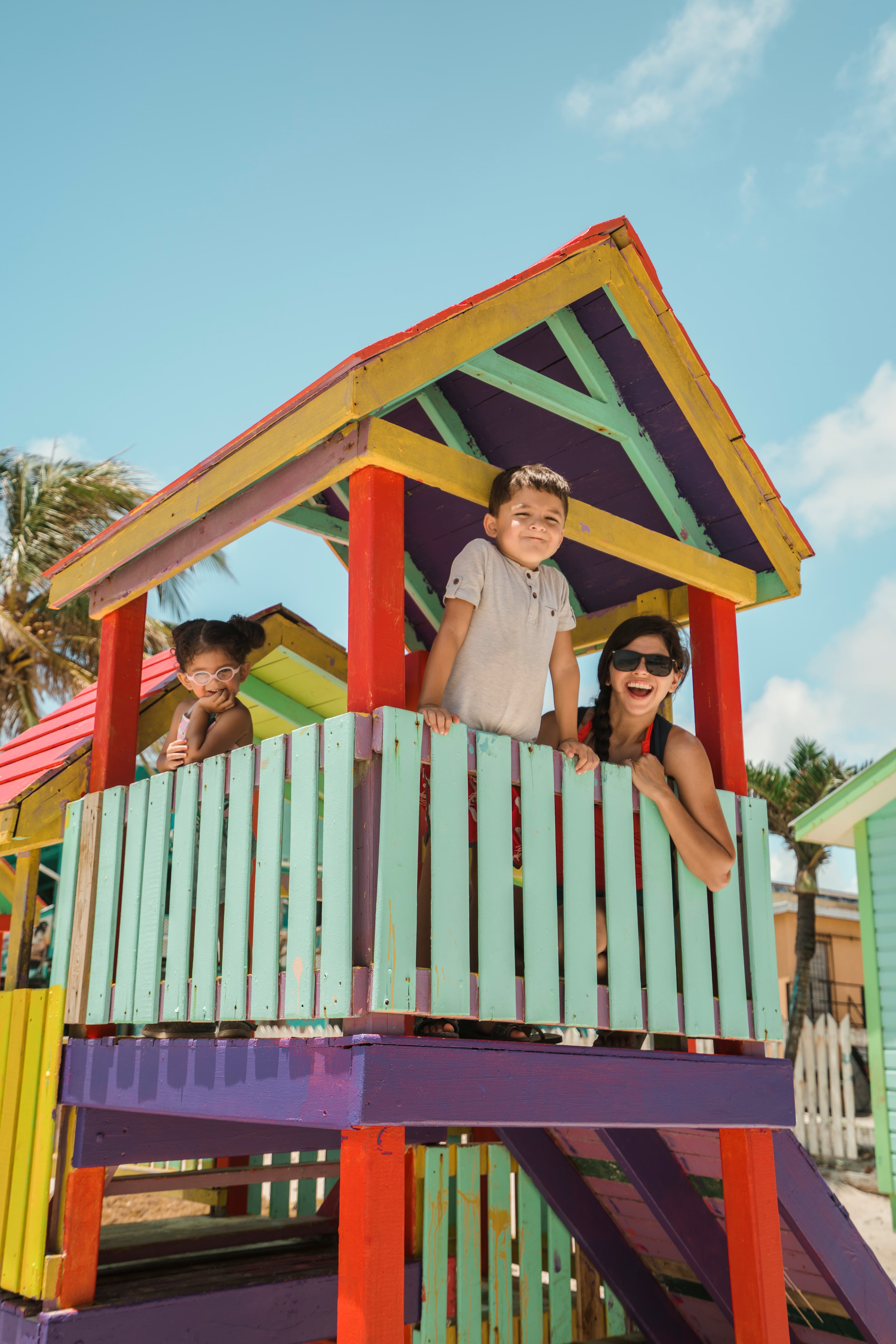 A woman and two children smile from a colorful play structure under a clear blue sky.