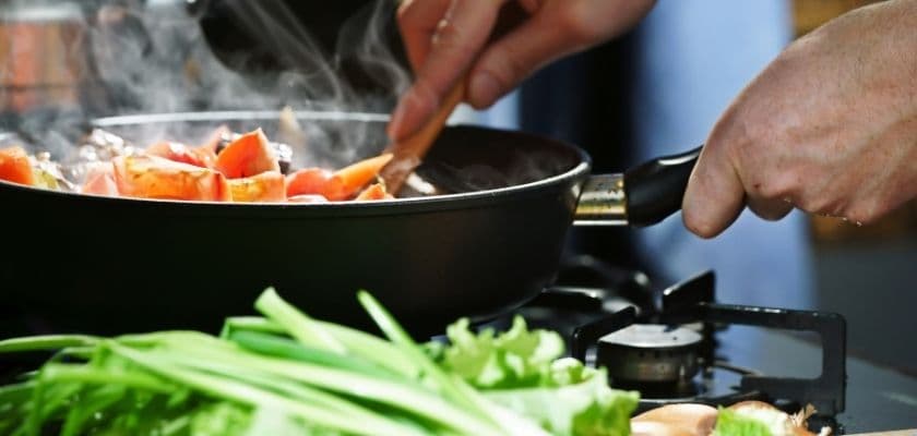 A person stirs chopped vegetables in a steaming frying pan on a stove, with fresh greens in the foreground.