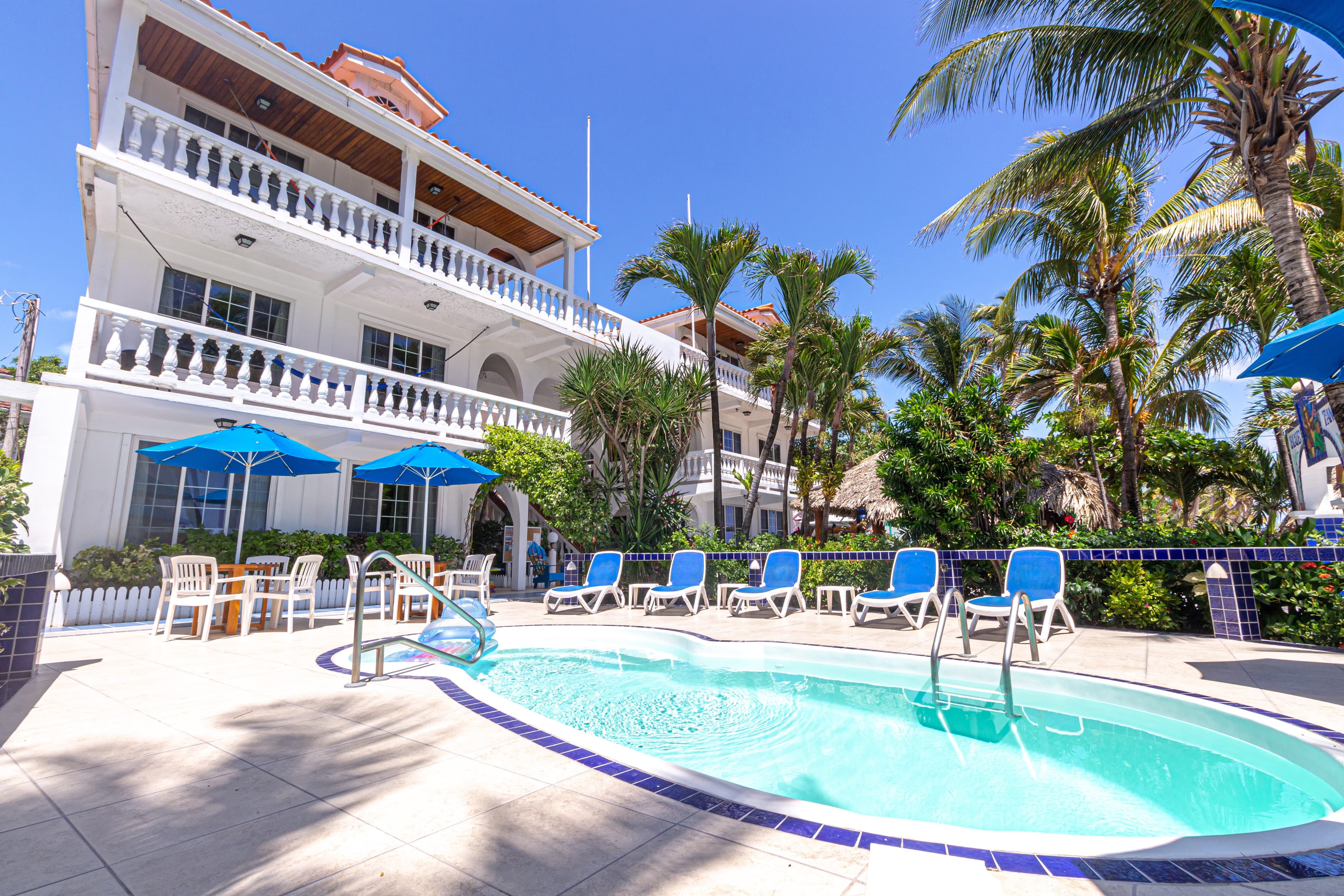 A sunny courtyard featuring a swimming pool, blue lounge chairs, and palm trees near a two-story white building.